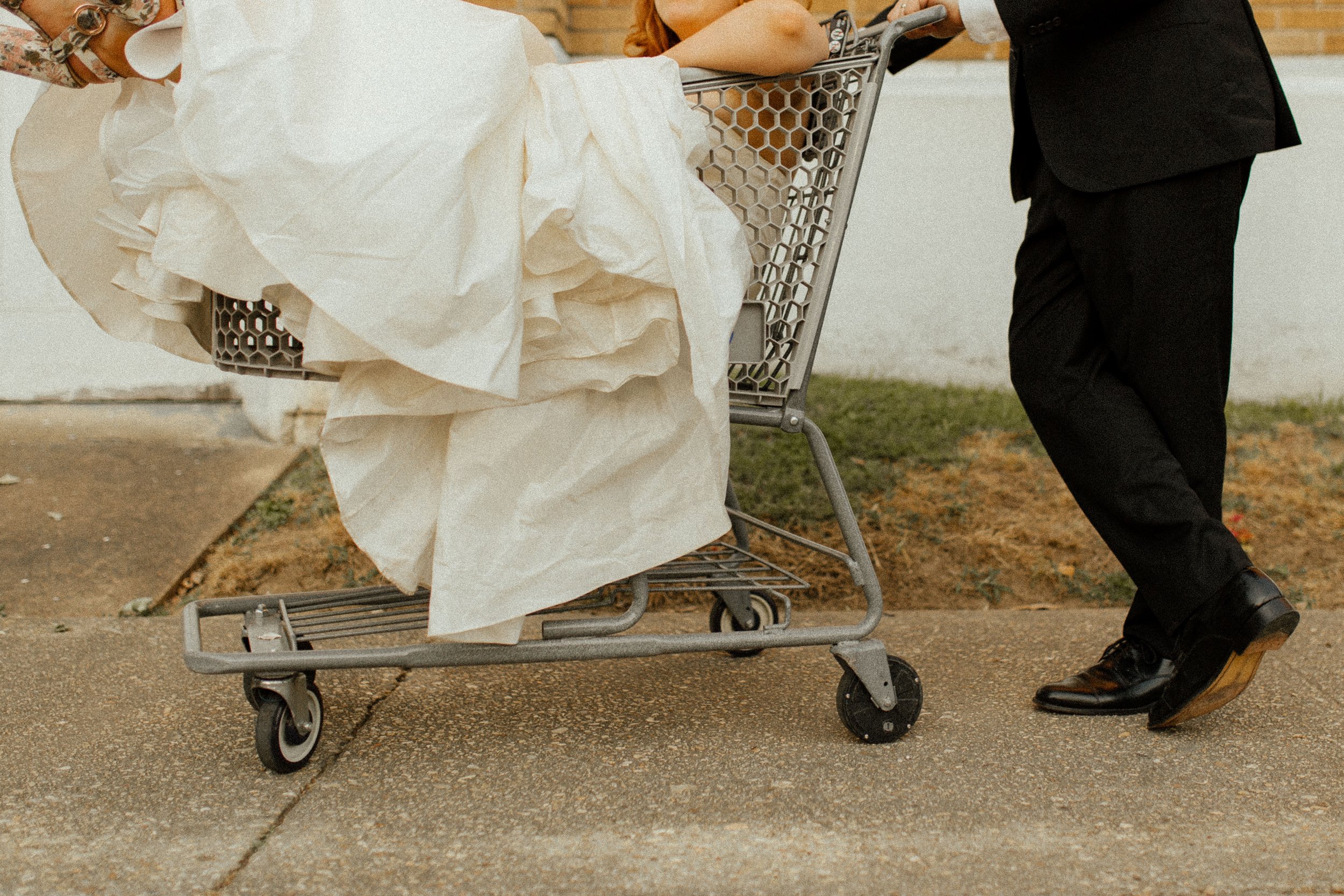 Groom pushes bride in a shopping cart along the sidewalk, her dress gathered around her in a fun and carefree pre wedding photos moment.