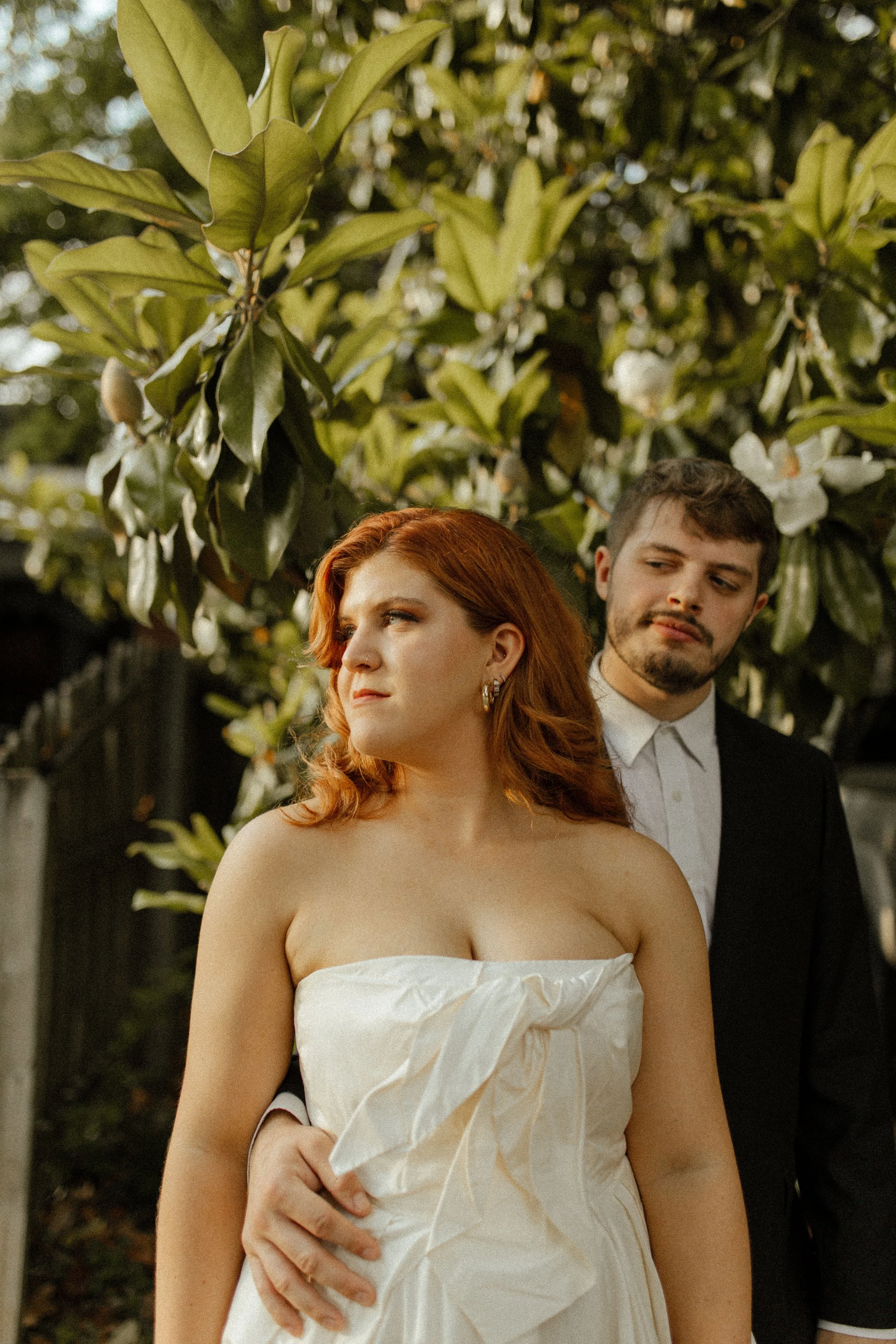 Bride stands in front of groom beneath magnolia leaves, both gazing off in different directions in a quiet, editorial moment.