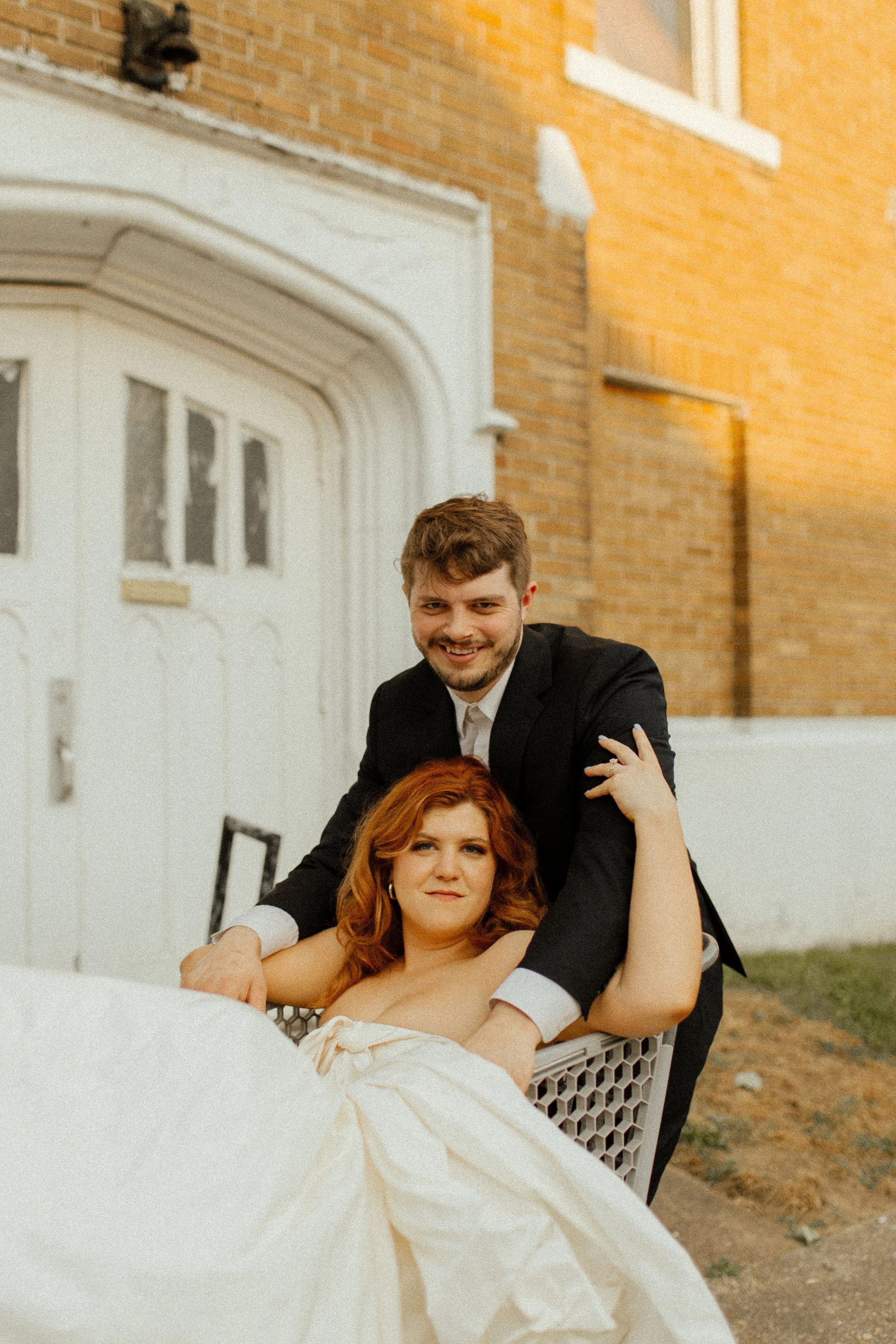 Groom leans over the back of a shopping cart as bride sits inside, both smiling in a playful and candid pre wedding photos scene.