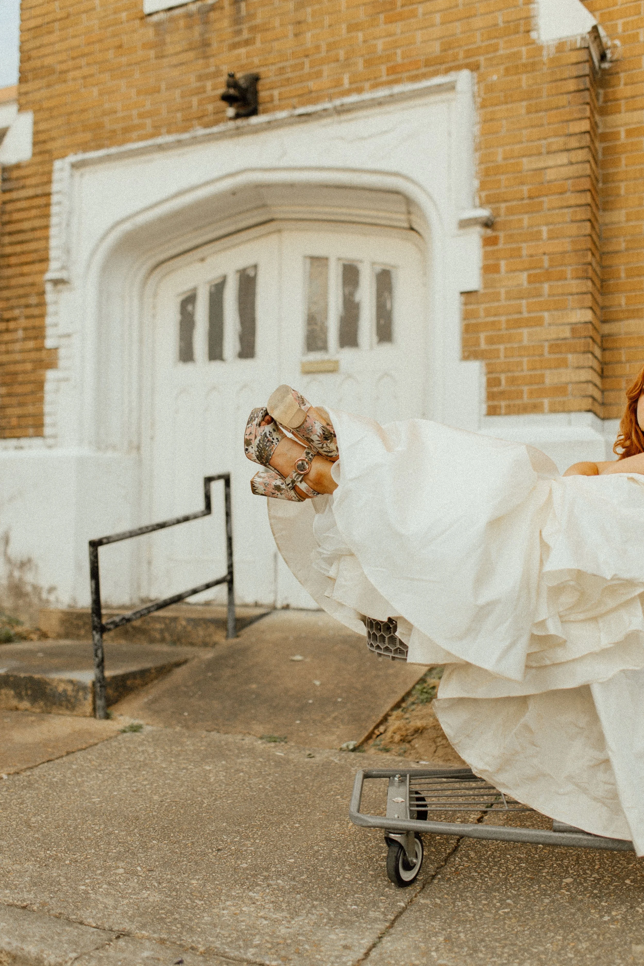 Bride lounges in a shopping cart with her dress spilling over the edge, boots peeking out in a playful pre wedding photos moment outside a brick building.