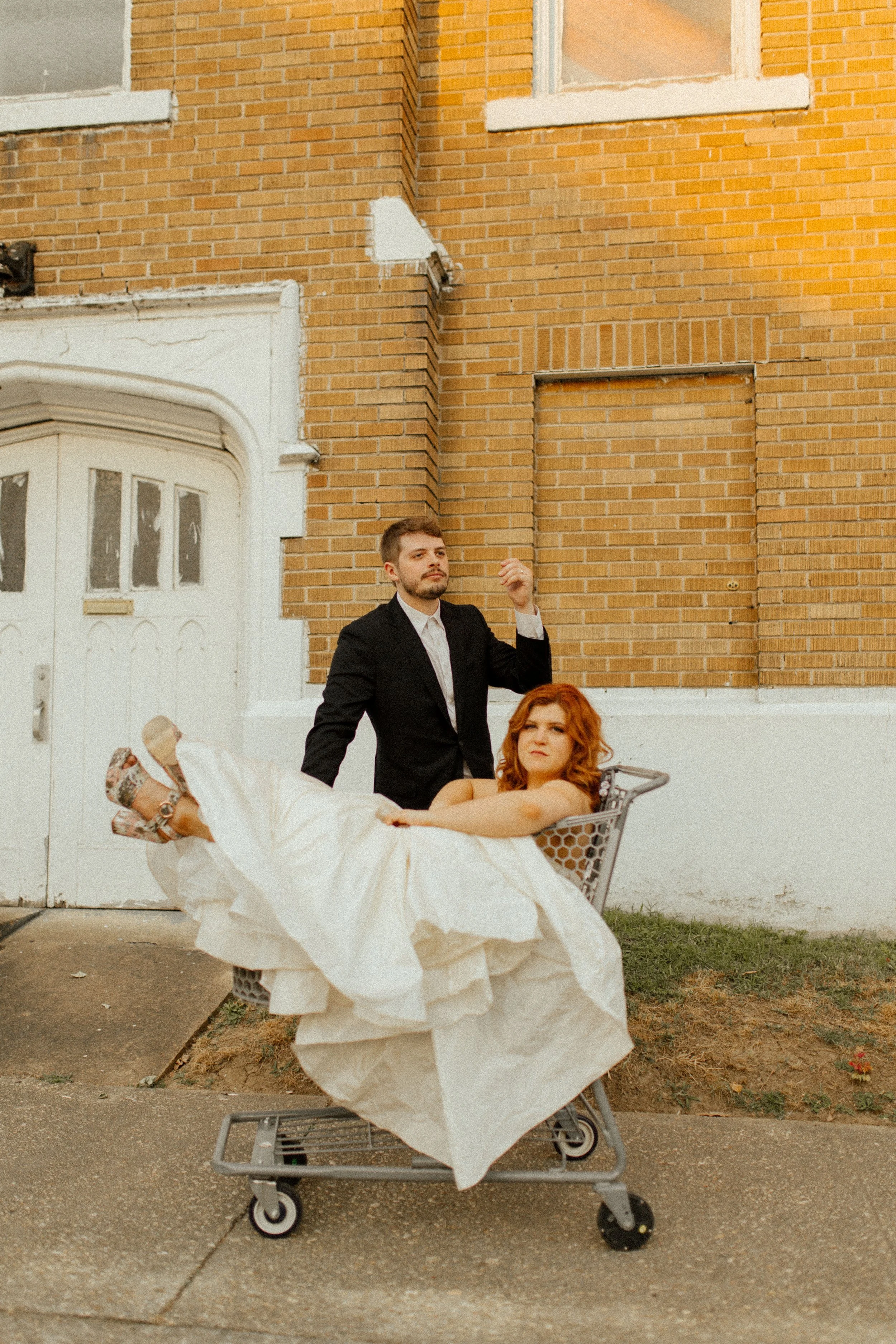 Bride lounges in a shopping cart while groom stands behind her outside a brick building, blending playful energy with editorial pre wedding photos style.