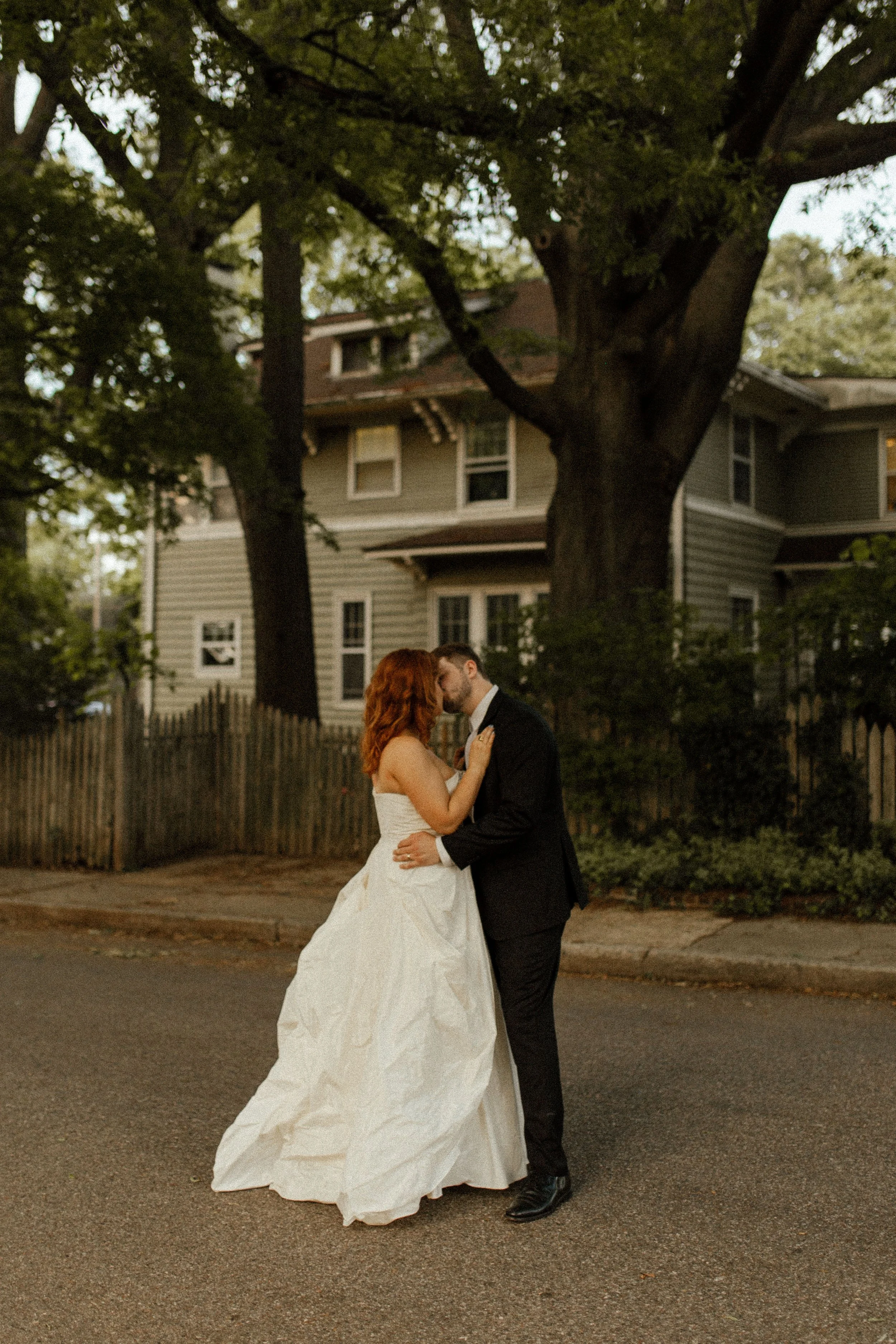 Couple shares a kiss in the middle of a quiet neighborhood street, surrounded by trees and warm evening light in romantic pre wedding photos.