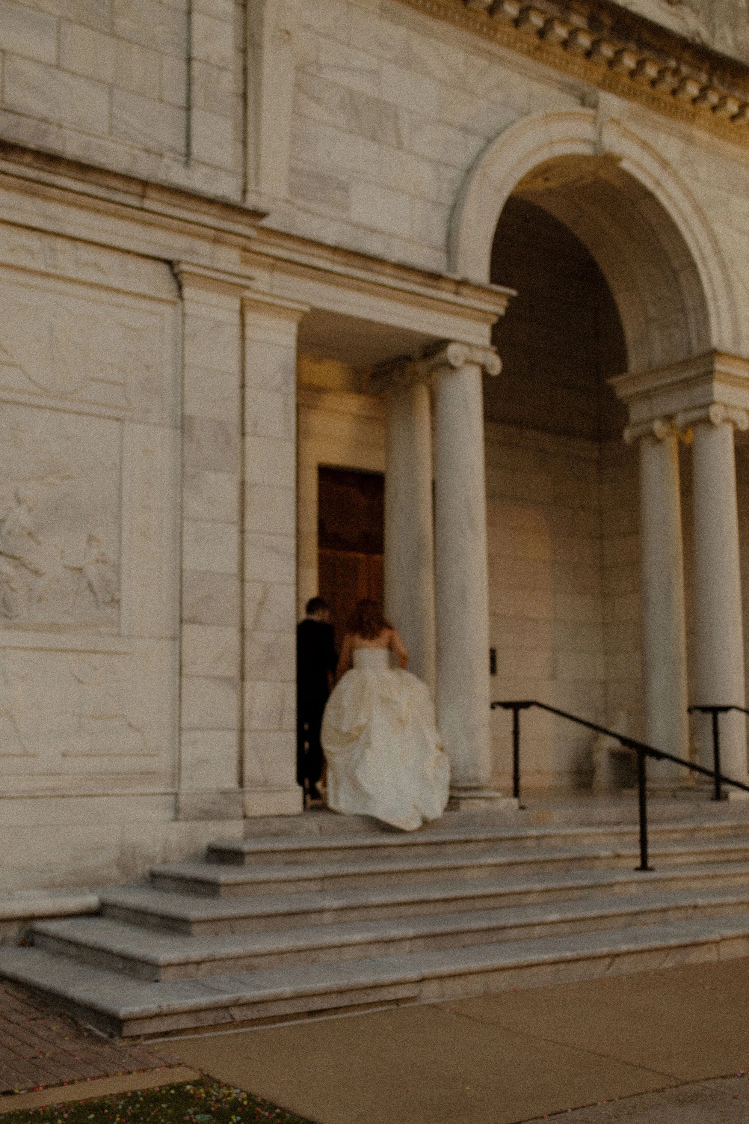Couple walks up grand stone steps toward an arched entrance, dress trailing behind in a cinematic and nostalgic pre wedding photos ending.