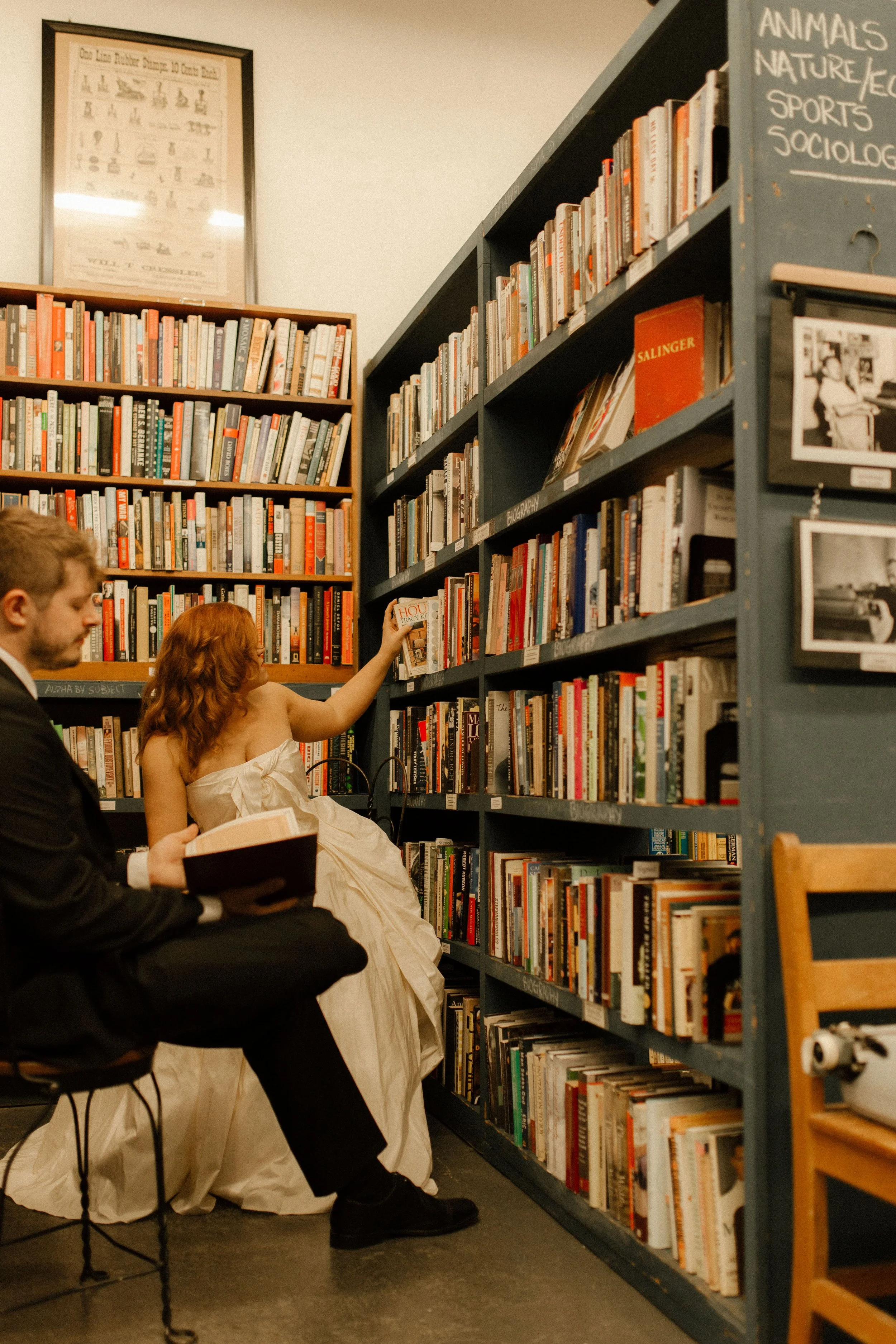 Bride reaches for a book on a high shelf while groom reads nearby, capturing a relaxed and candid bookstore moment.