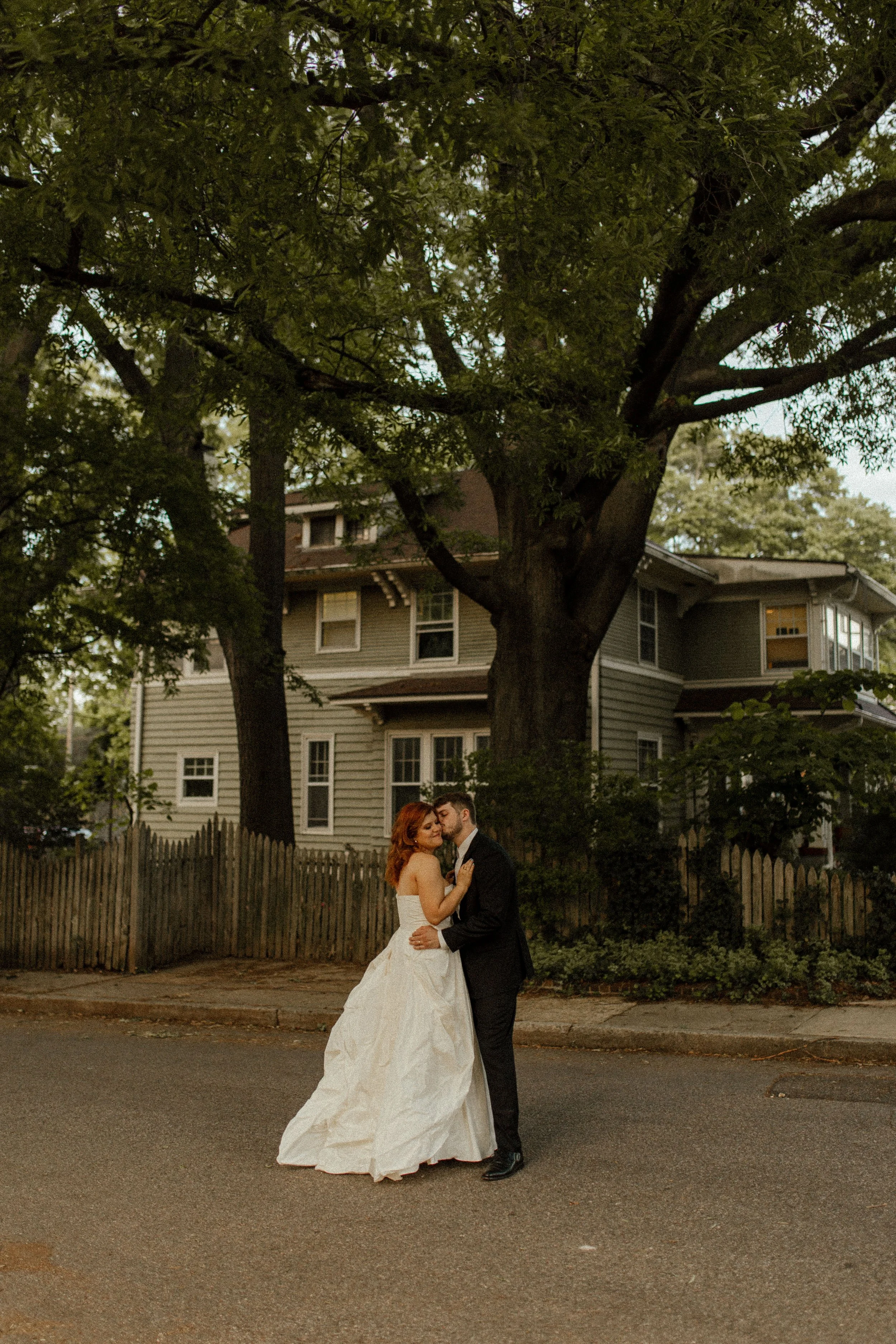 Groom wraps his arms around bride outside a brick building, both smiling softly in a warm and affectionate pre wedding photos scene.