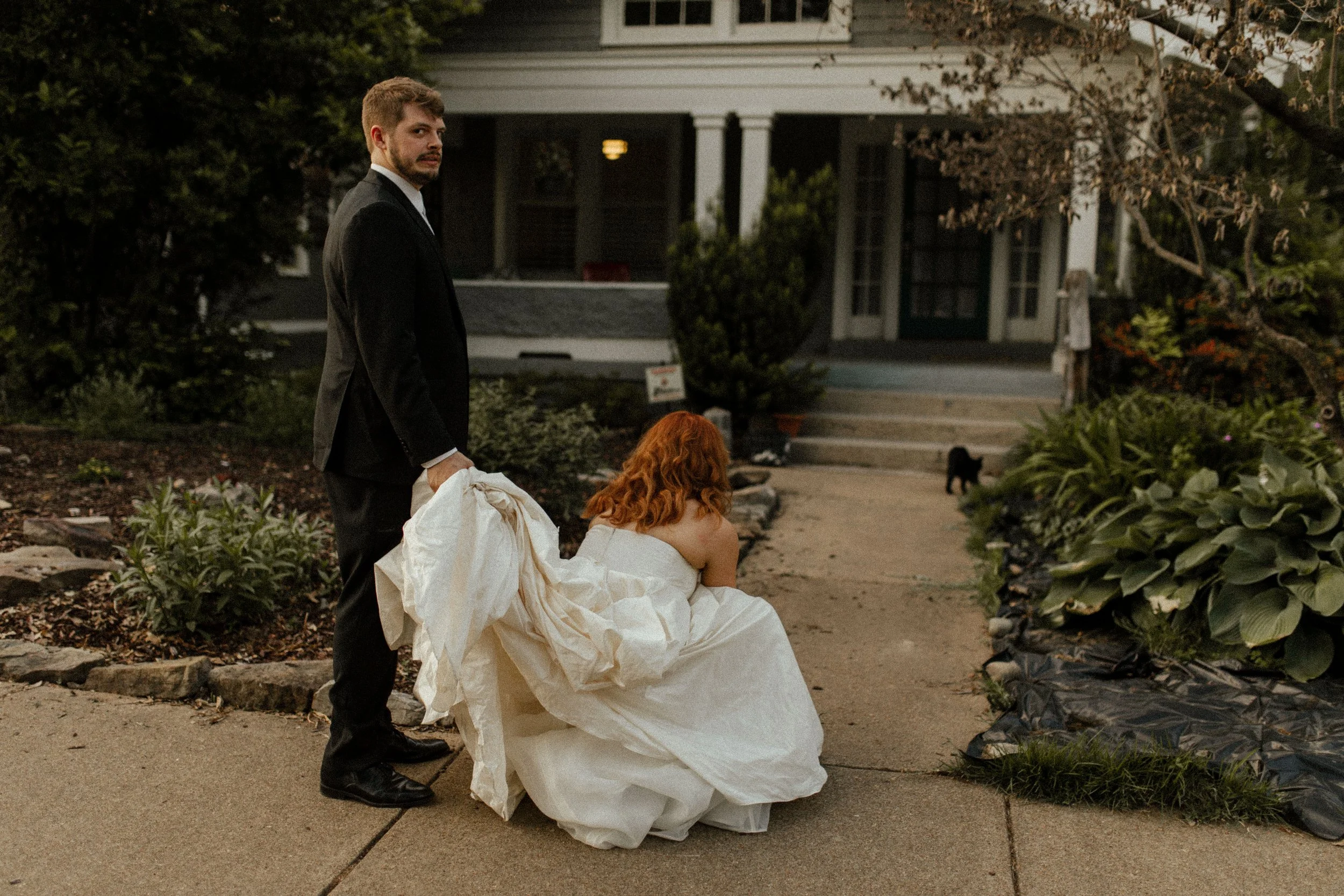 Groom holds the bride’s dress as she crouches down on a garden path near a house, capturing a candid and playful pre wedding photos moment.