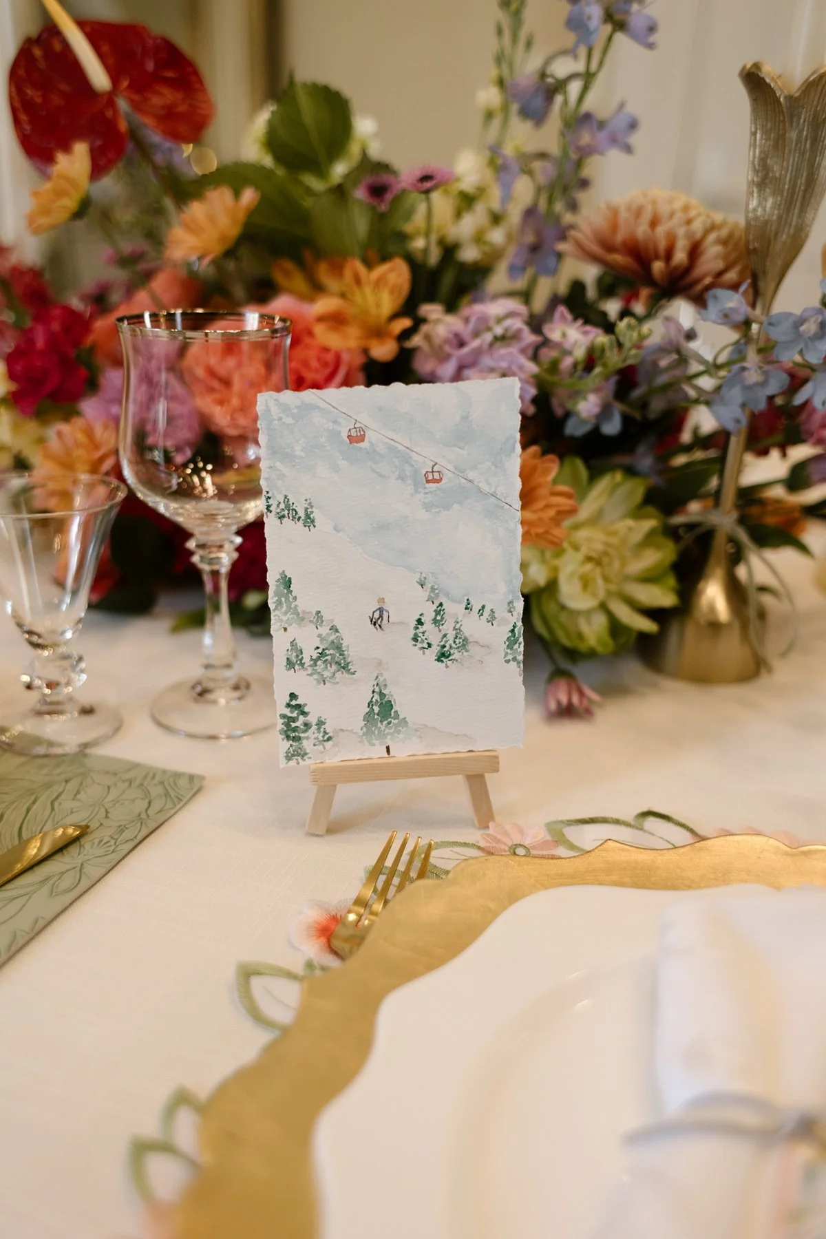 hand-painted mountain scene table number displayed among colorful floral arrangements and vintage glassware