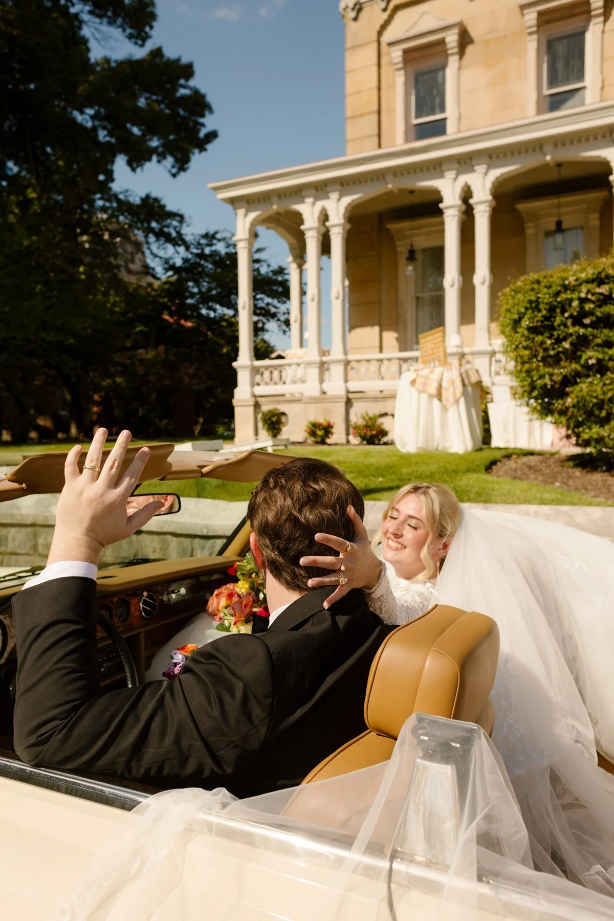 newlyweds driving away in vintage convertible in front of historic memphis home, veil flowing in the wind