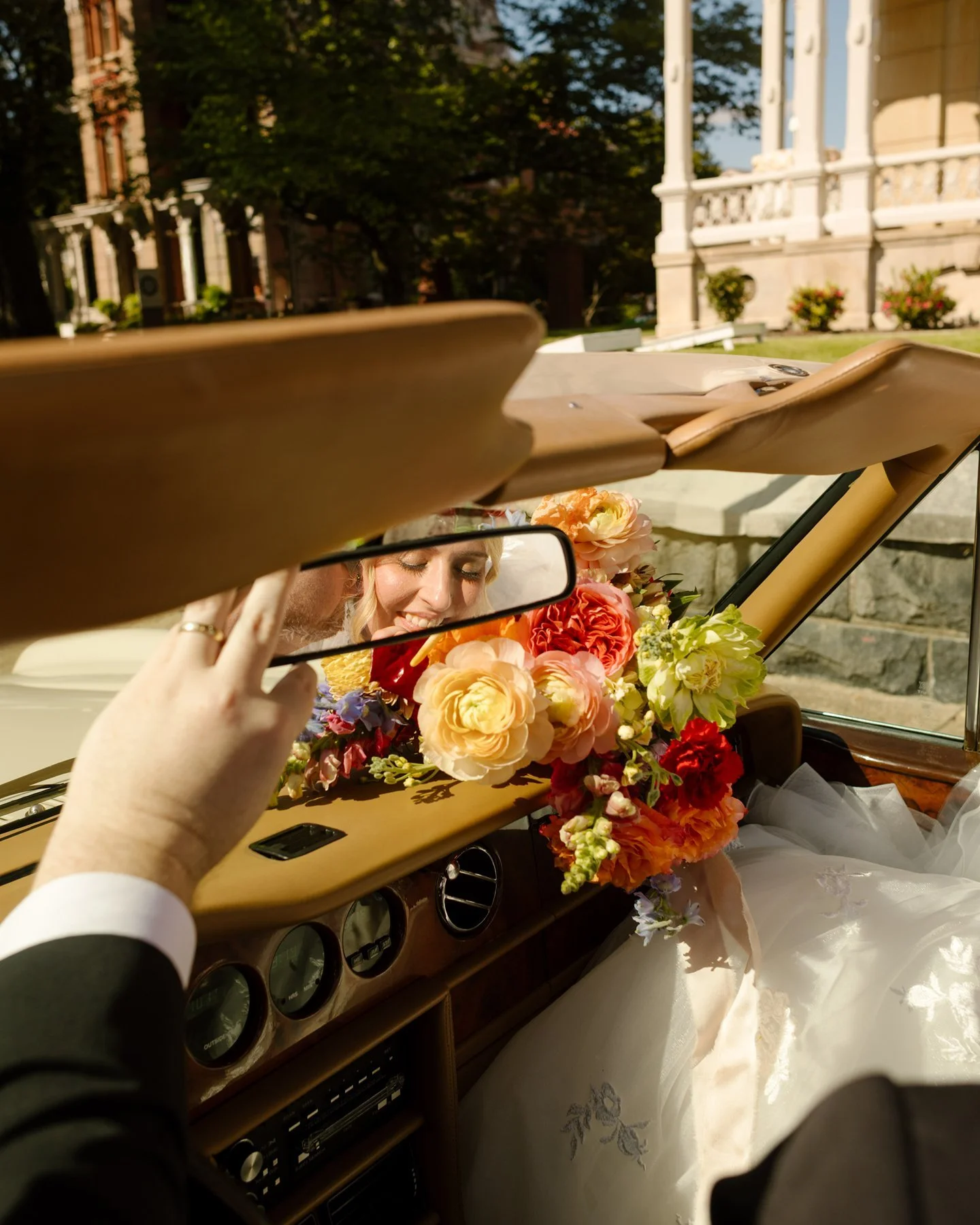 reflection of bride smiling in car mirror surrounded by vibrant bouquet, candid and playful just-married moment