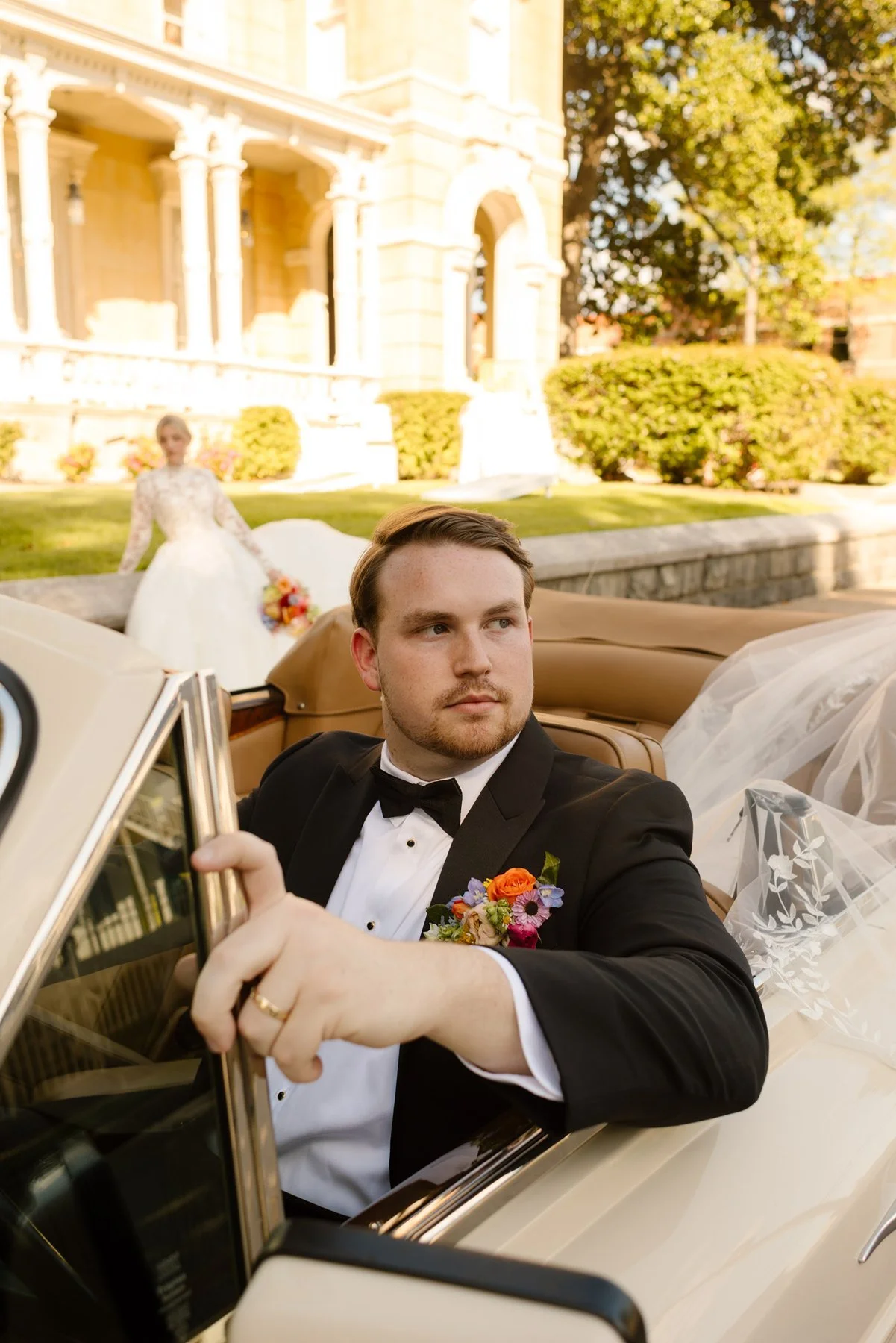 groom in tux sitting in vintage convertible with bride in background, editorial wedding portrait with classic styling
