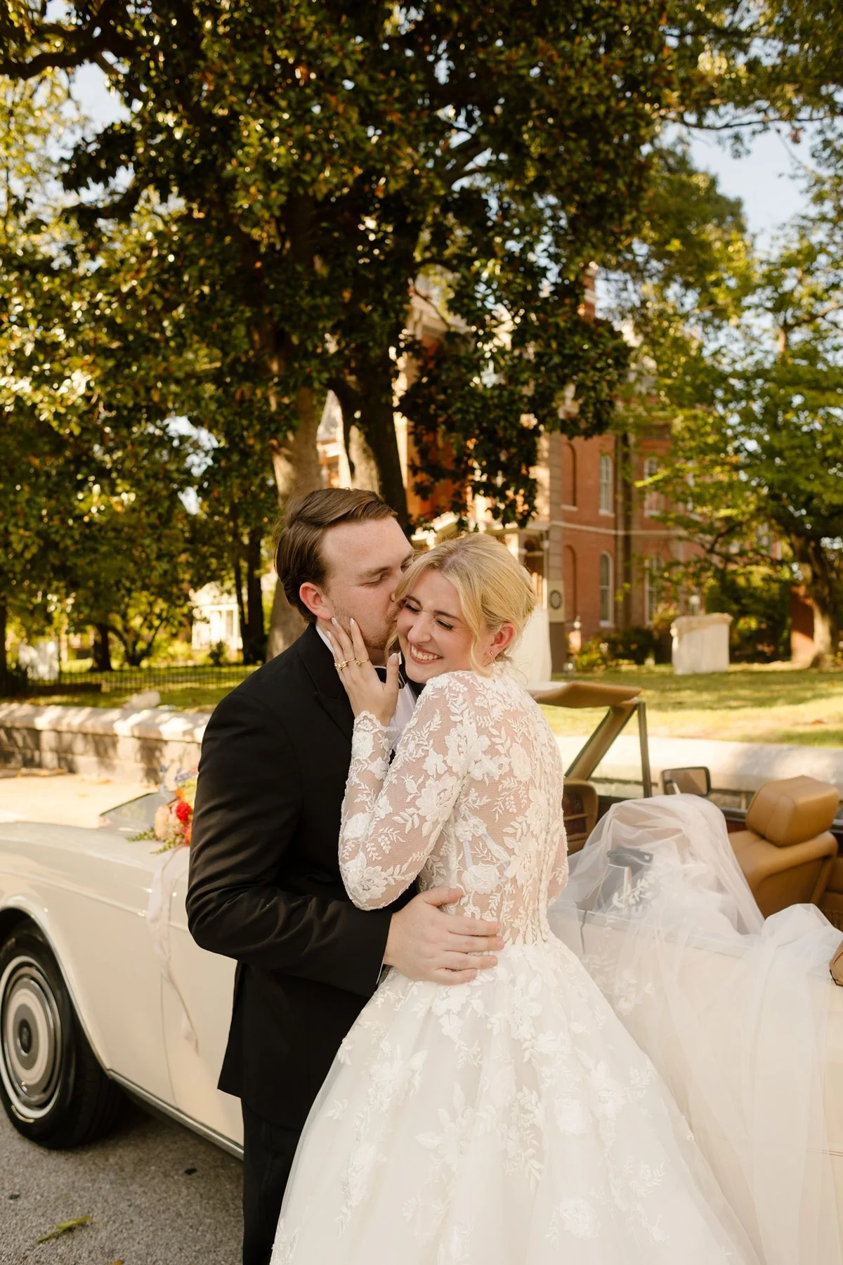 groom kissing bride’s cheek beside vintage car, joyful and affectionate newlywed moment