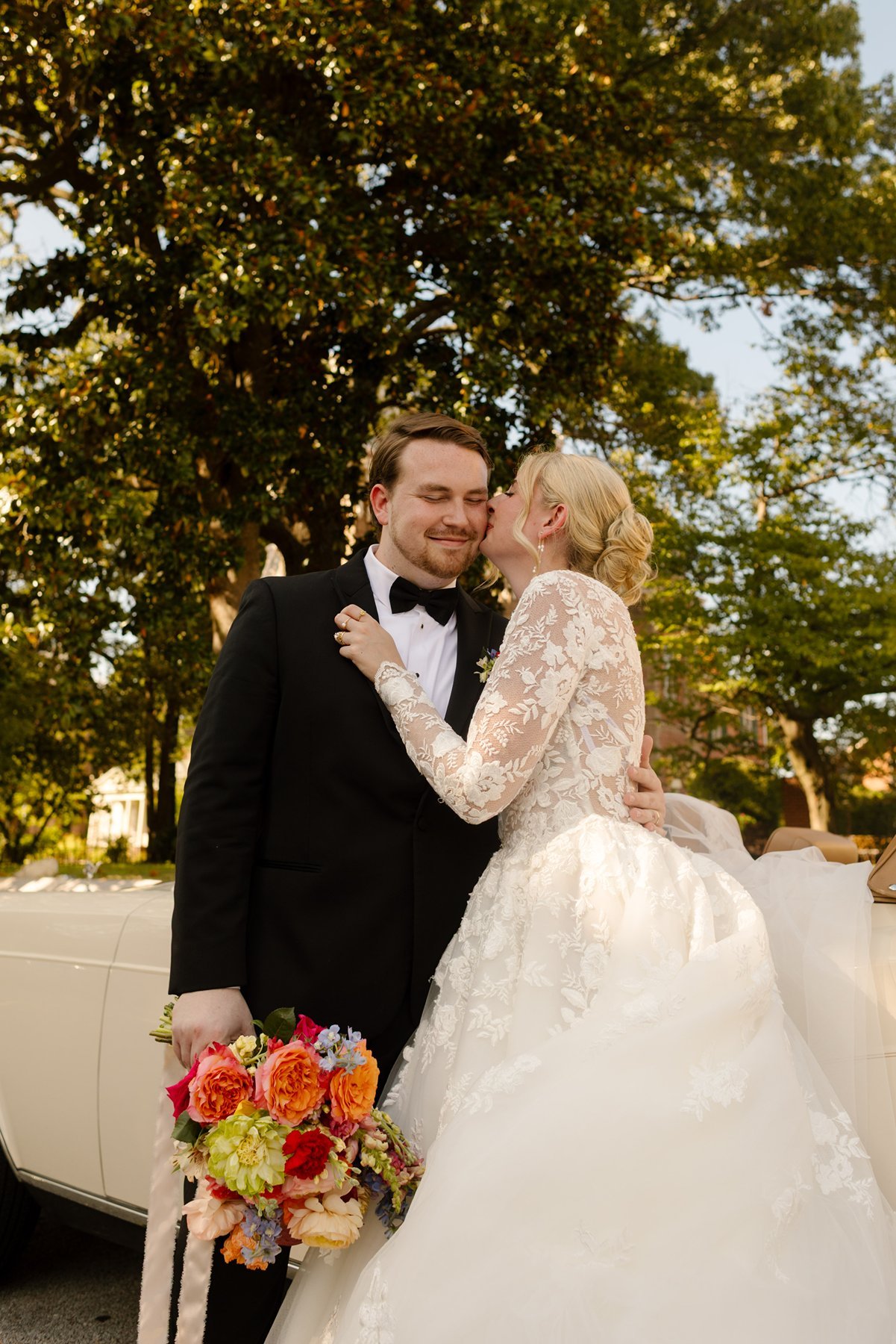 Bride kissing groom on the cheek while he holds a bright bouquet, standing beside a vintage car.