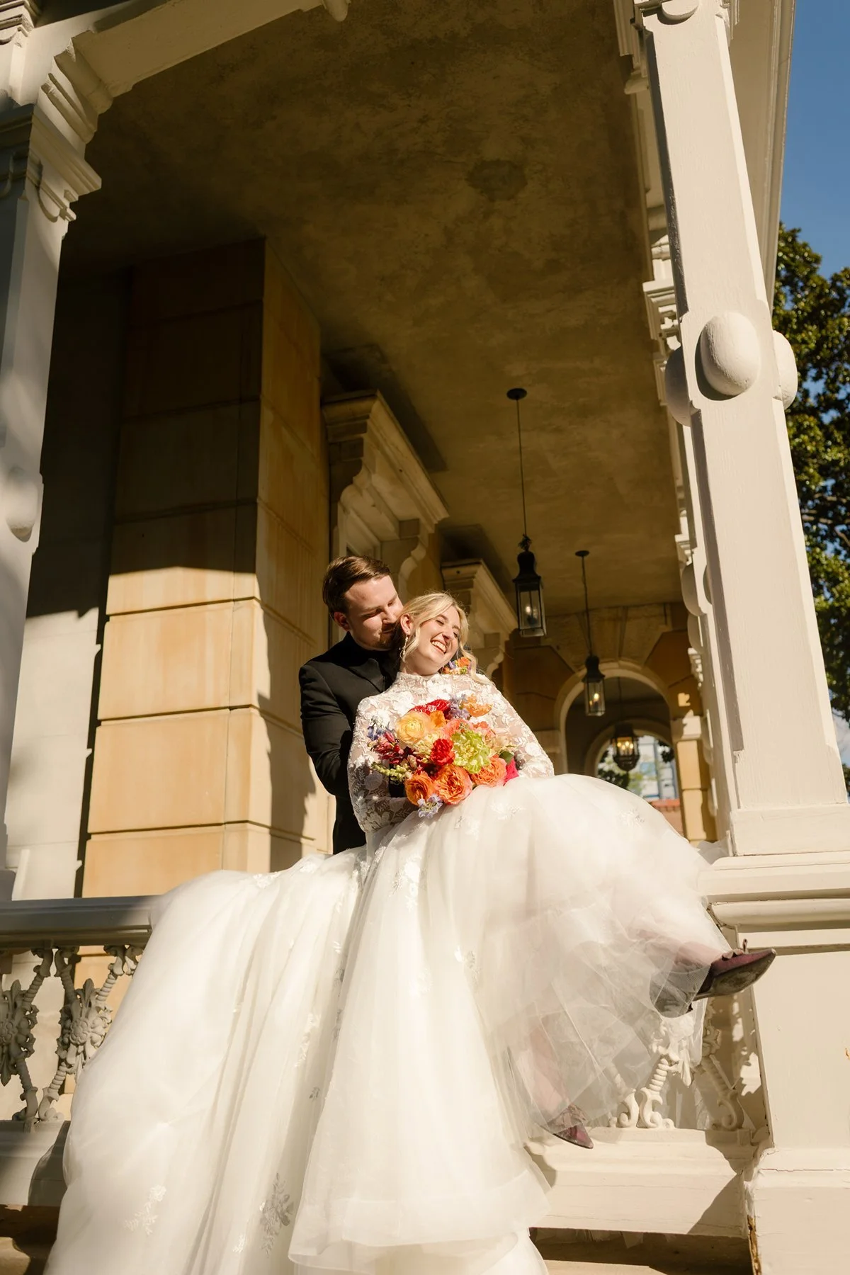 Groom standing behind bride on a porch, holding her as she leans back laughing with bouquet in hand.
