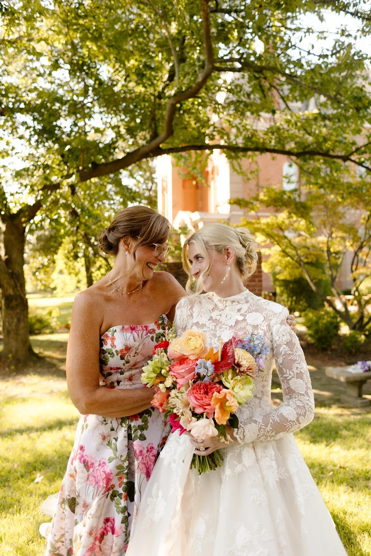 Bride and an older woman in a floral dress smiling at each other, holding a colorful bouquet under dappled sunlight.