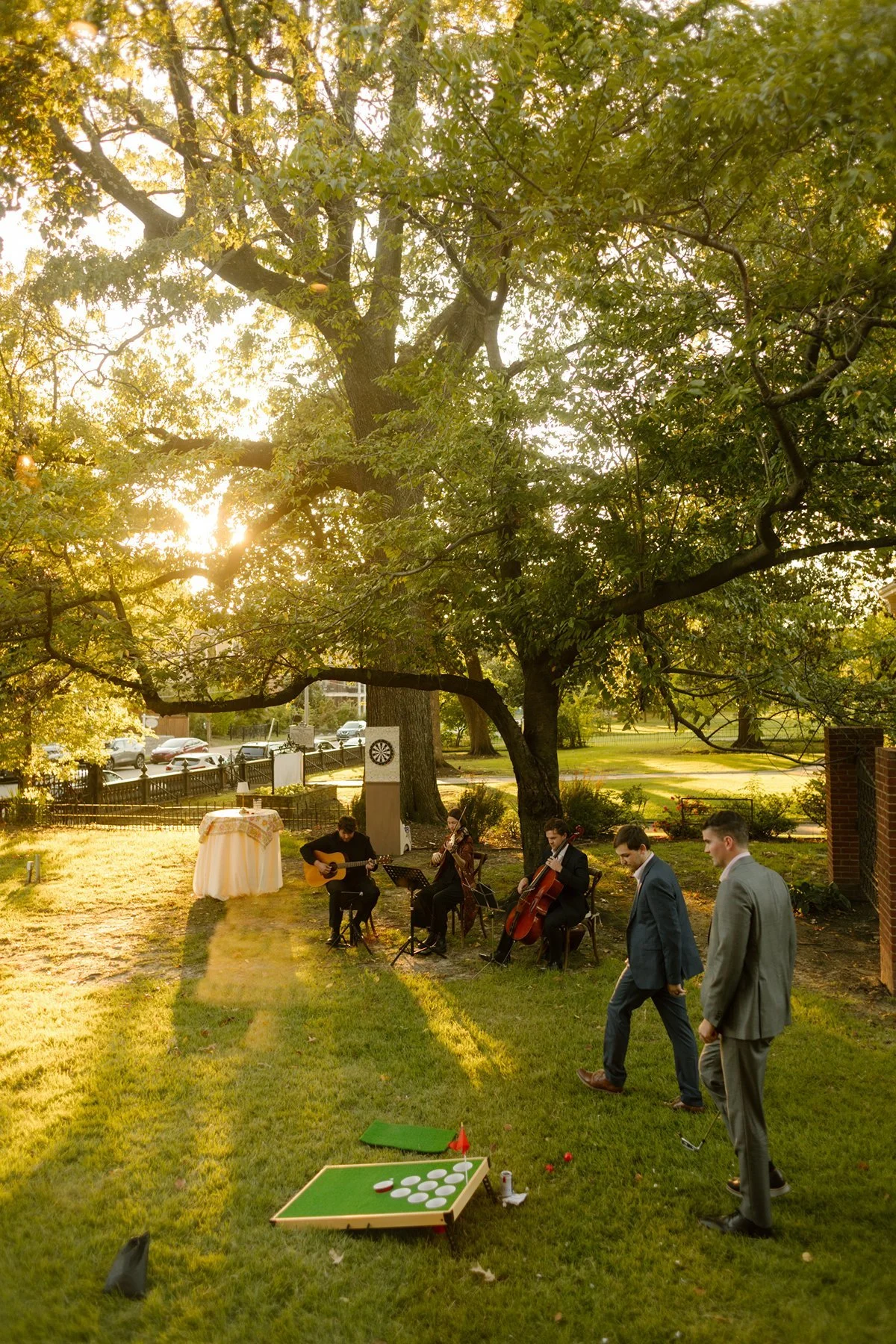 String quartet playing outdoors under trees during golden hour while guests mingle and lawn games sit nearby.