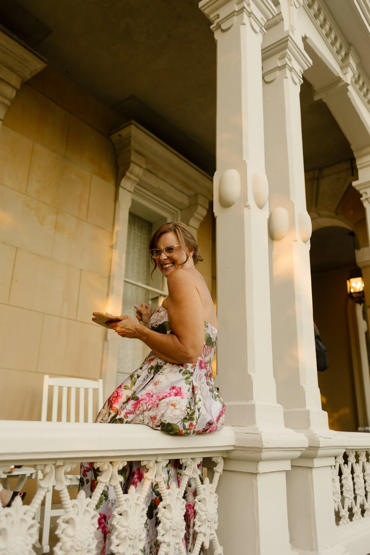 Woman in a floral dress sitting on a porch railing, smiling while holding a small plate.