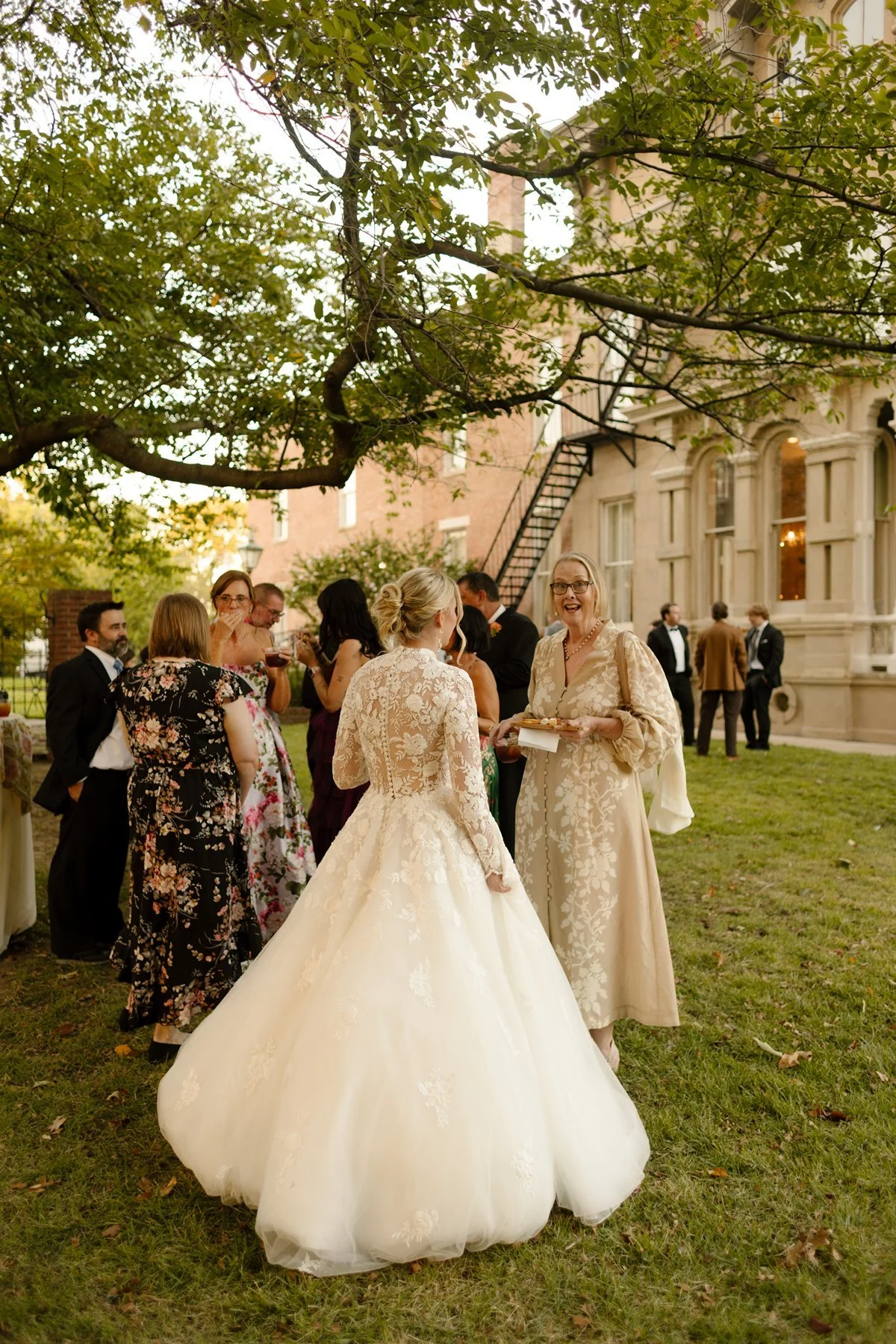 Bride in a lace gown greeting guests on a lawn during cocktail hour, surrounded by trees and a historic building.