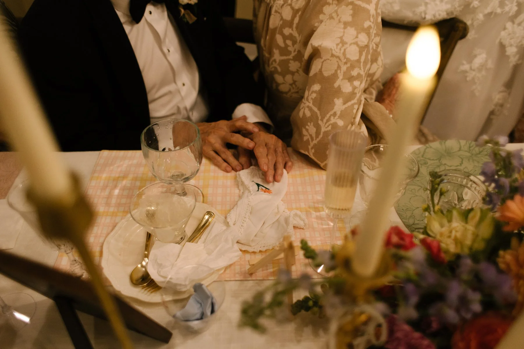Bride and groom seated close together at their reception table, hands resting together beside candlelight and soft details.