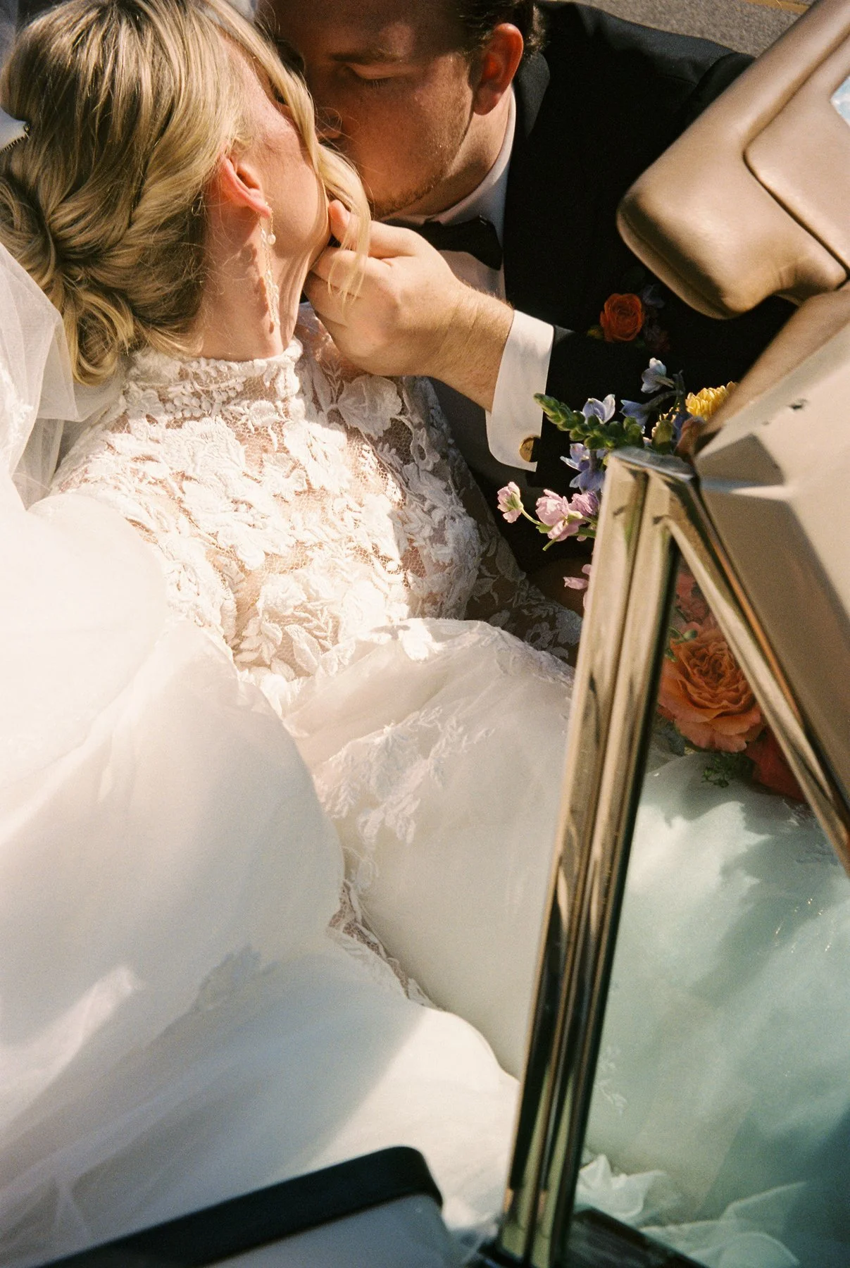 Bride and groom kissing in the front seat of a vintage car, her tulle dress and bouquet tucked around them in sunlight.