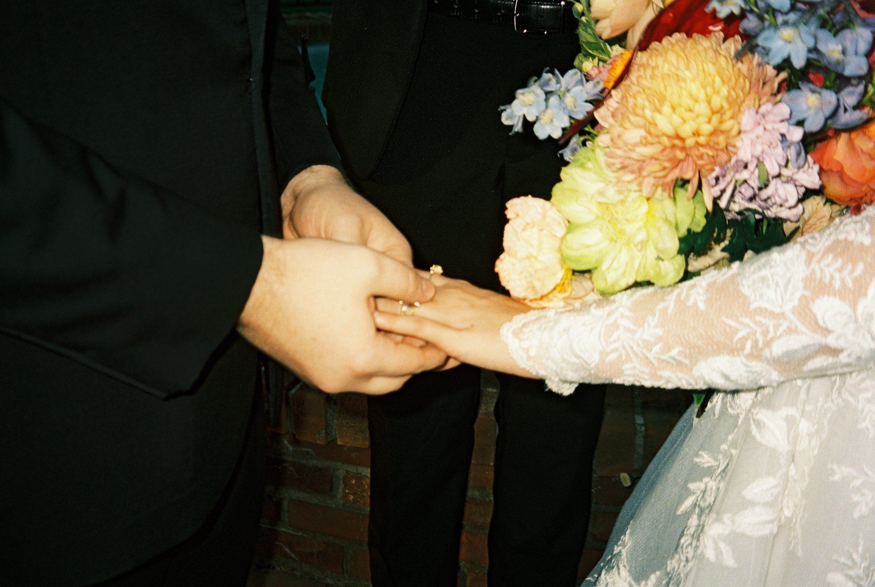 Close-up of the groom sliding a ring onto the bride’s finger, her lace sleeve and colorful bouquet visible beside their hands.