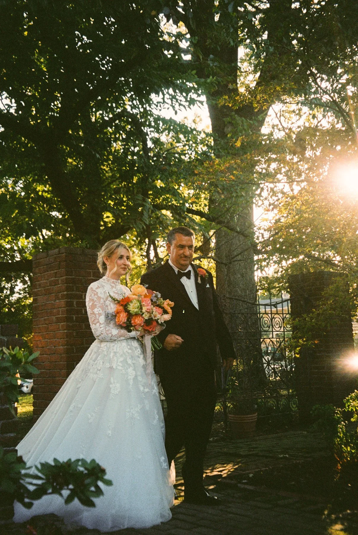 Bride walking arm in arm with an older man down a garden path, holding a colorful bouquet as sunlight filters through the trees.
