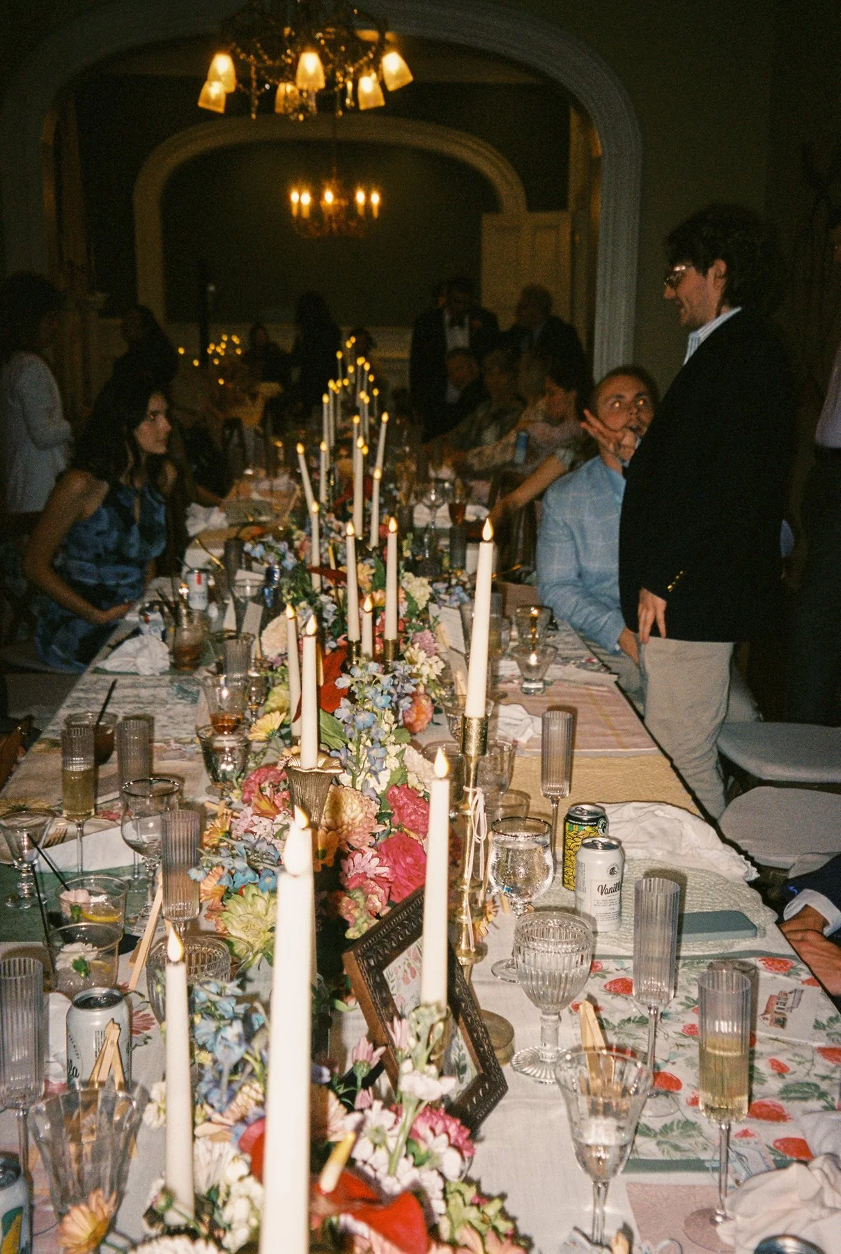 Long candlelit reception table filled with florals, glassware, and guests mid-conversation in a dim, elegant dining room.