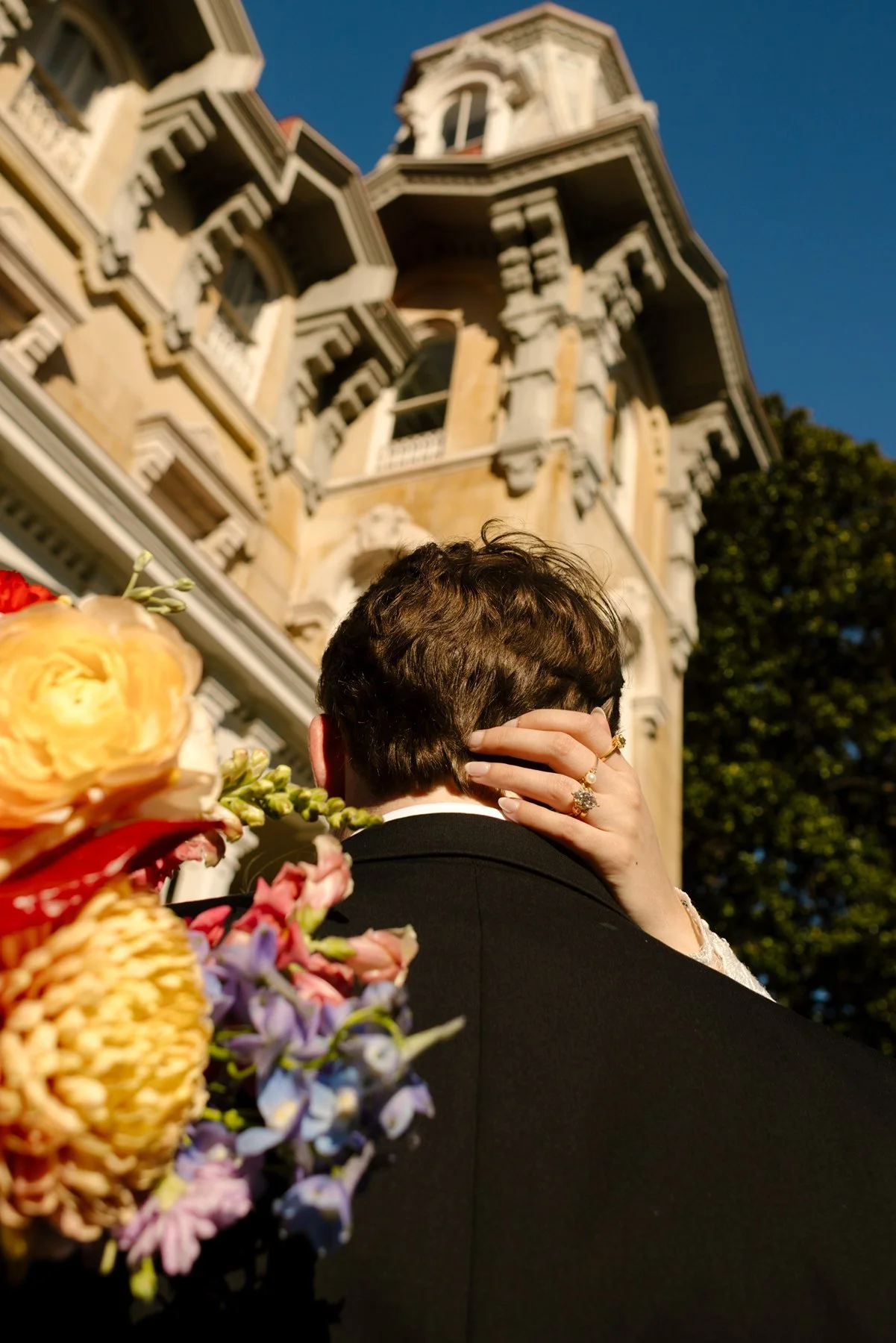 Close-up from behind the groom as the bride wraps her hand around his neck, her ring and bouquet visible against a grand historic building.