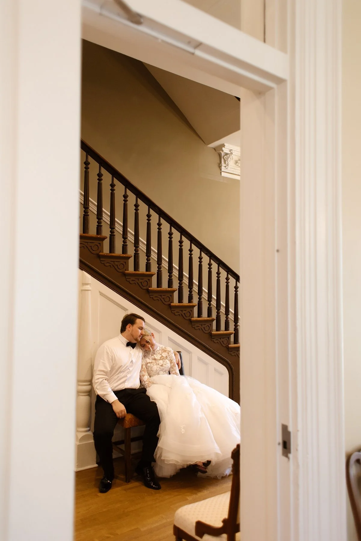 Bride and groom resting together on a bench by a staircase indoors, leaning into each other in a quiet moment.