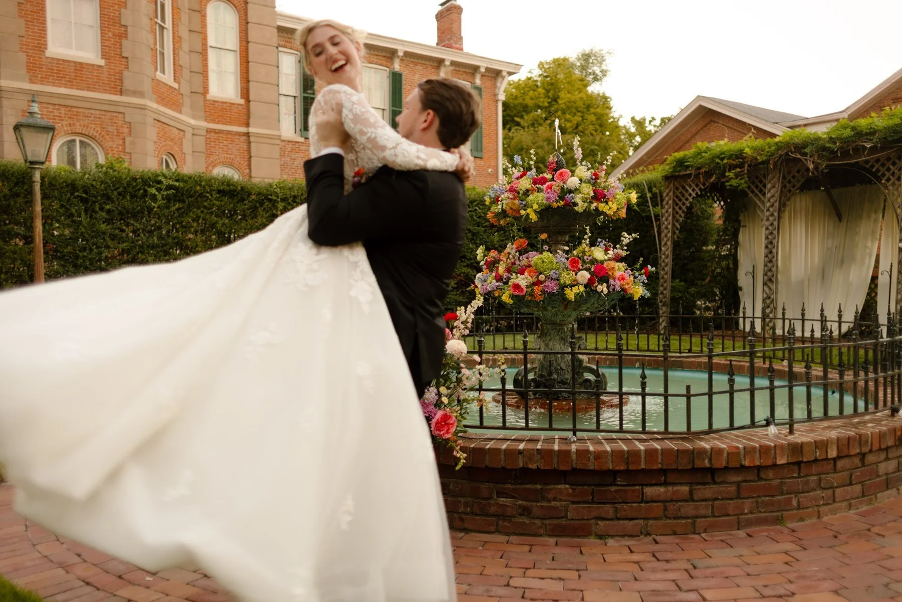 Groom lifting the bride as she laughs, her dress flowing beside a flower-filled fountain in a garden courtyard.