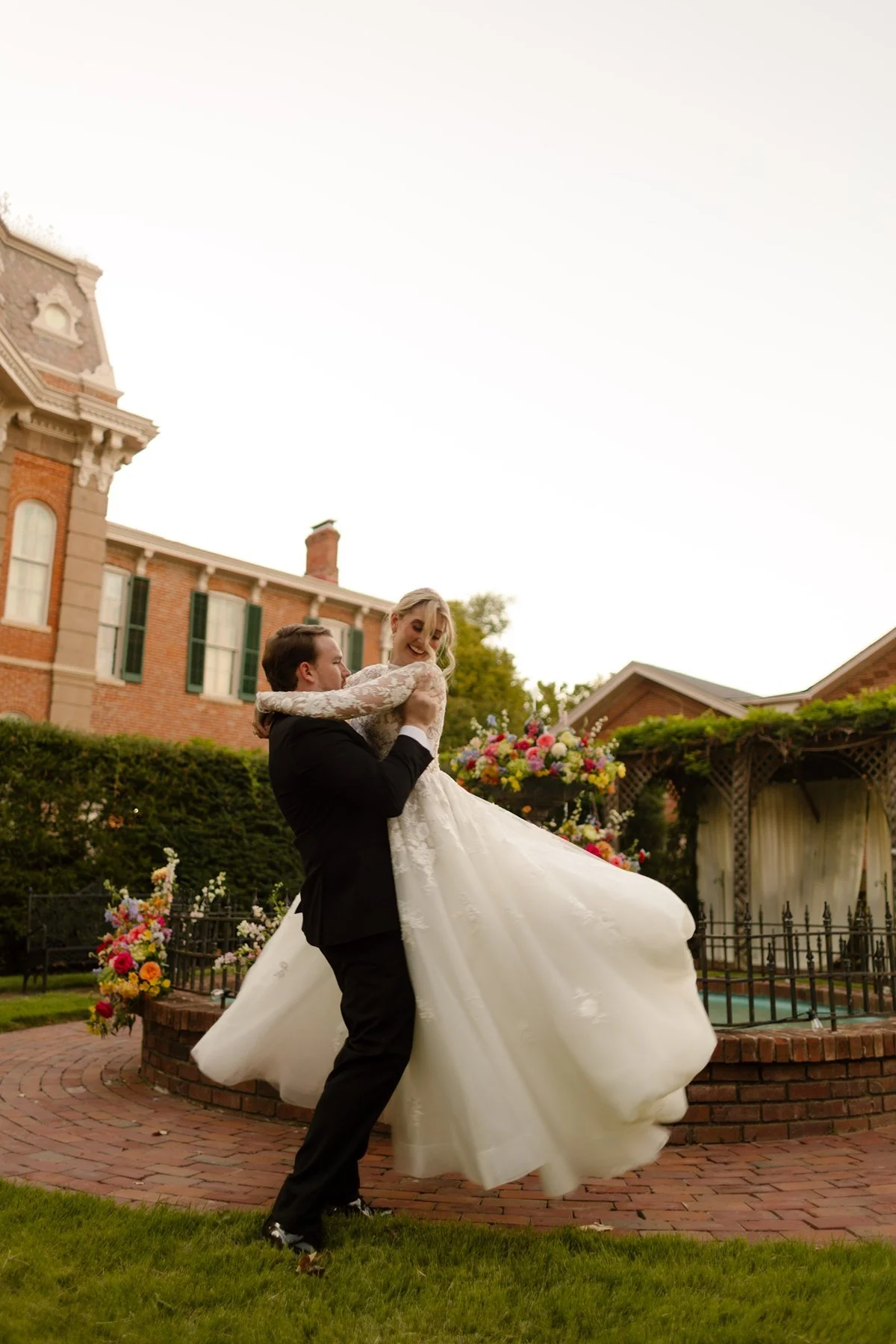 Groom lifting the bride as her dress swirls around them in a garden courtyard by a fountain.