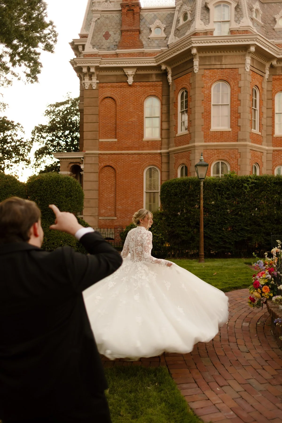Bride twirling away as the groom points toward her, playful moment in front of a historic brick estate.
