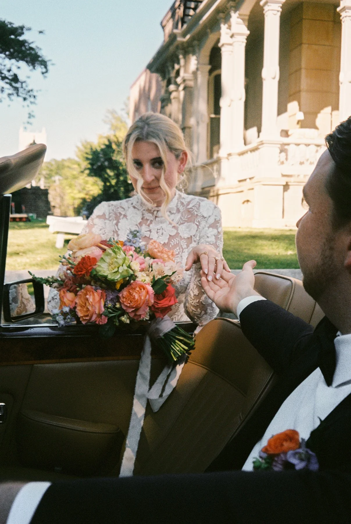 Bride standing outside a vintage car, holding her bouquet while reaching for the groom’s hand inside.