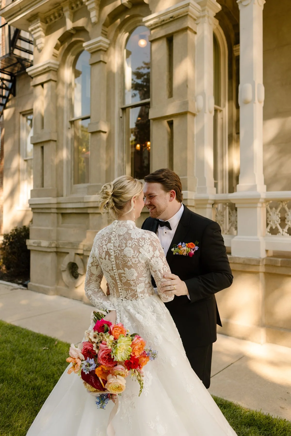 Bride and groom smiling at each other in front of a historic building, her bouquet held behind her back.