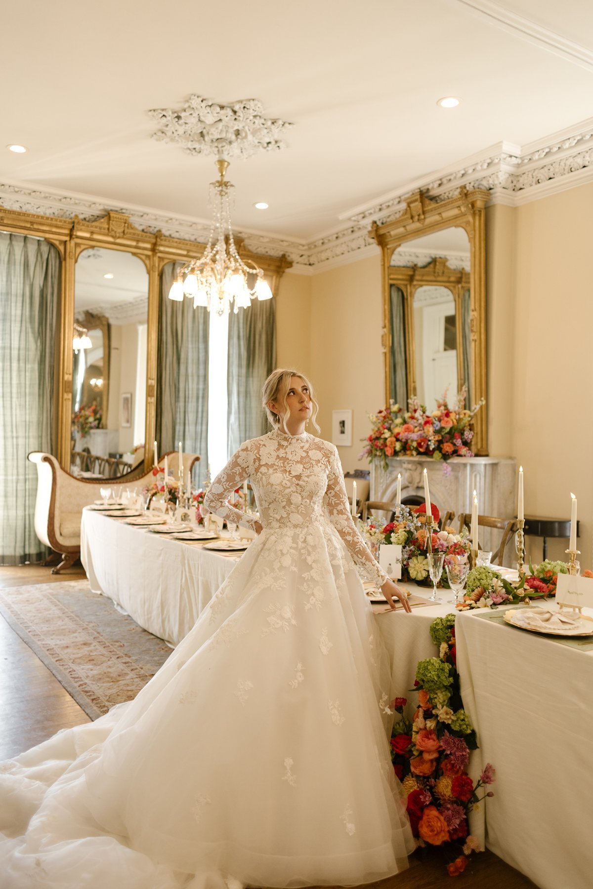 Bride standing beside a long, candlelit reception table in an ornate room, looking up beneath a chandelier.
