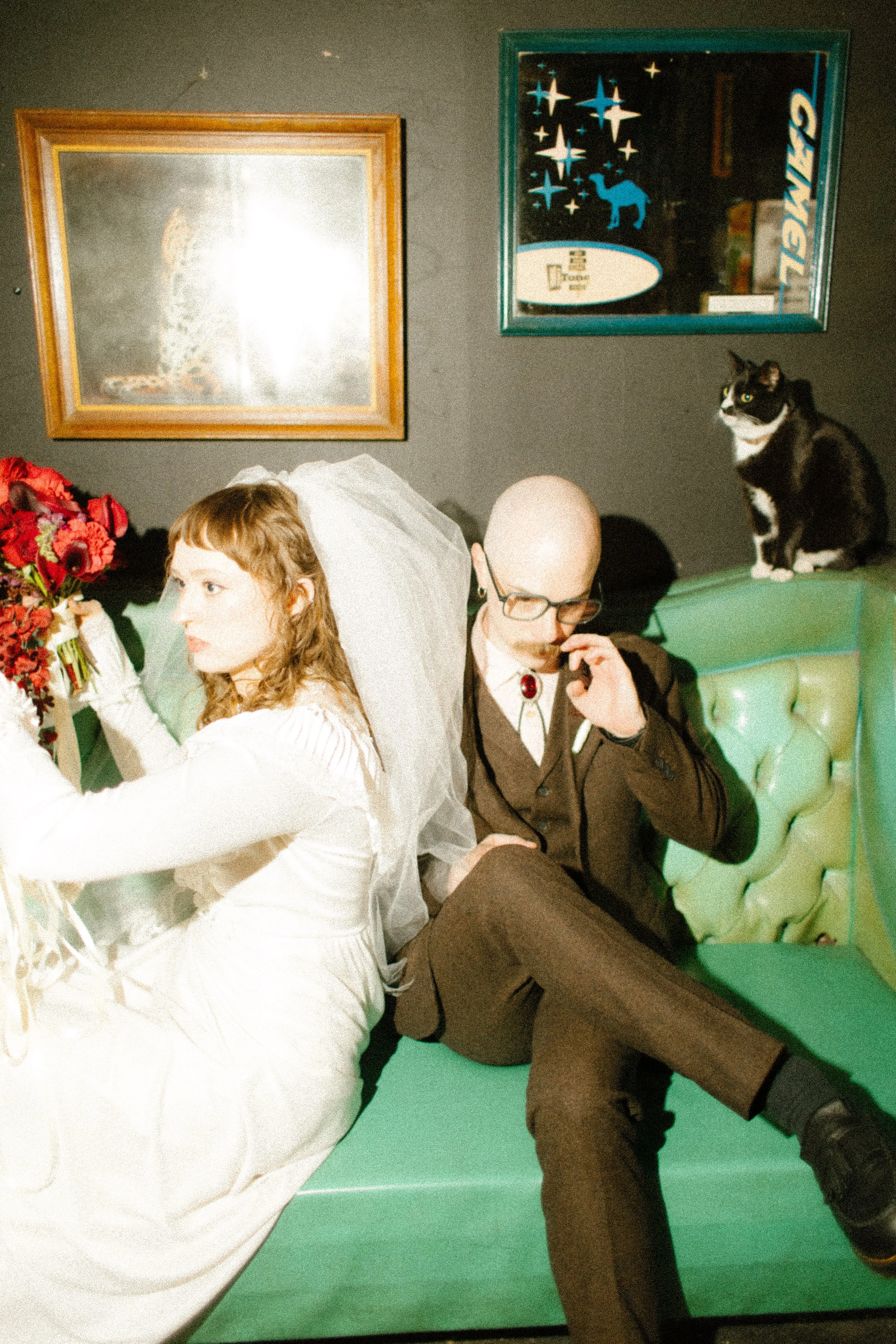 Bride and groom posing on a couch with a cat nearby in a moody bar setting, capturing quirky and authentic wedding moments