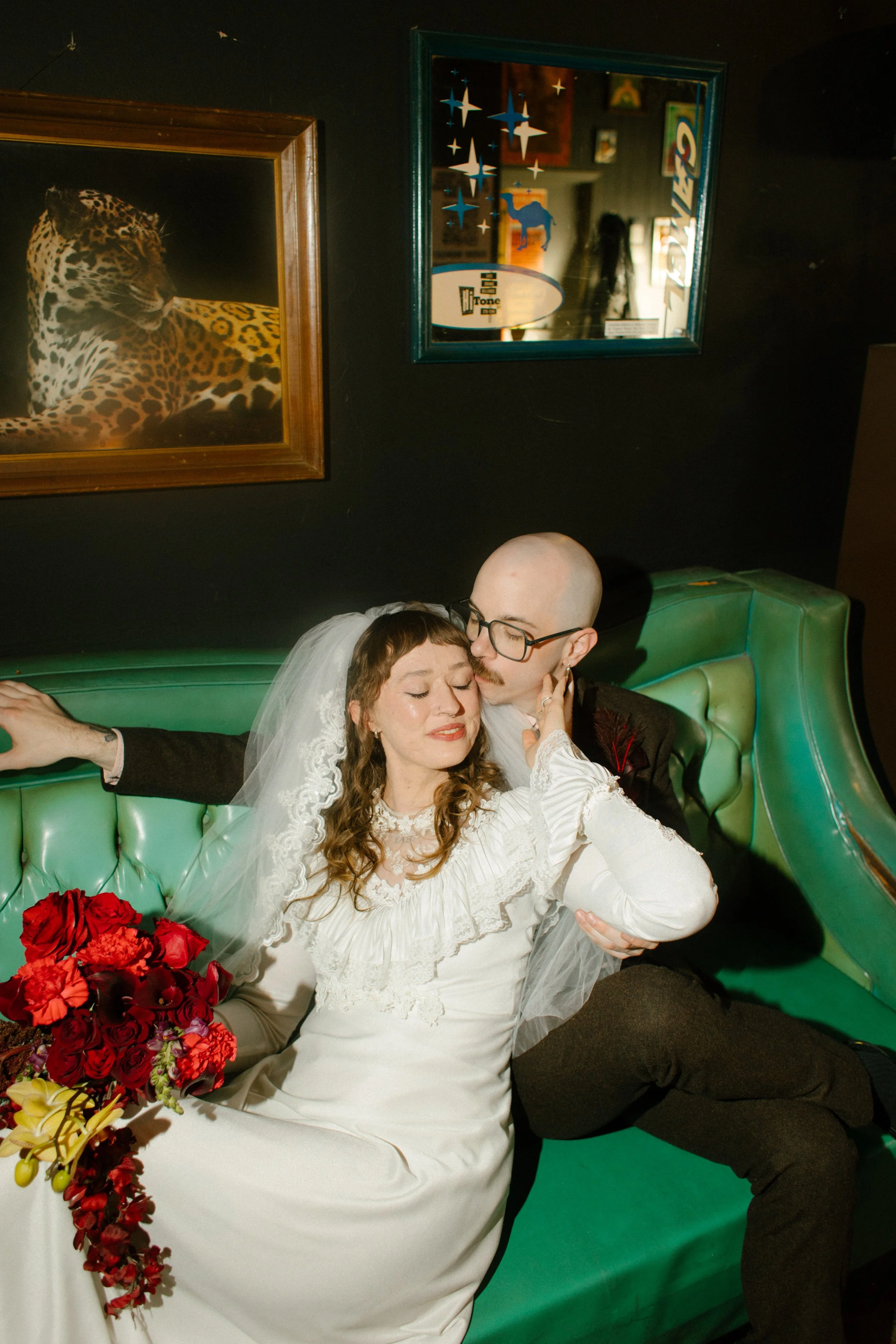 Bride and groom sitting together on a green couch in a dimly lit bar, sharing an intimate moment during their non-traditional wedding day