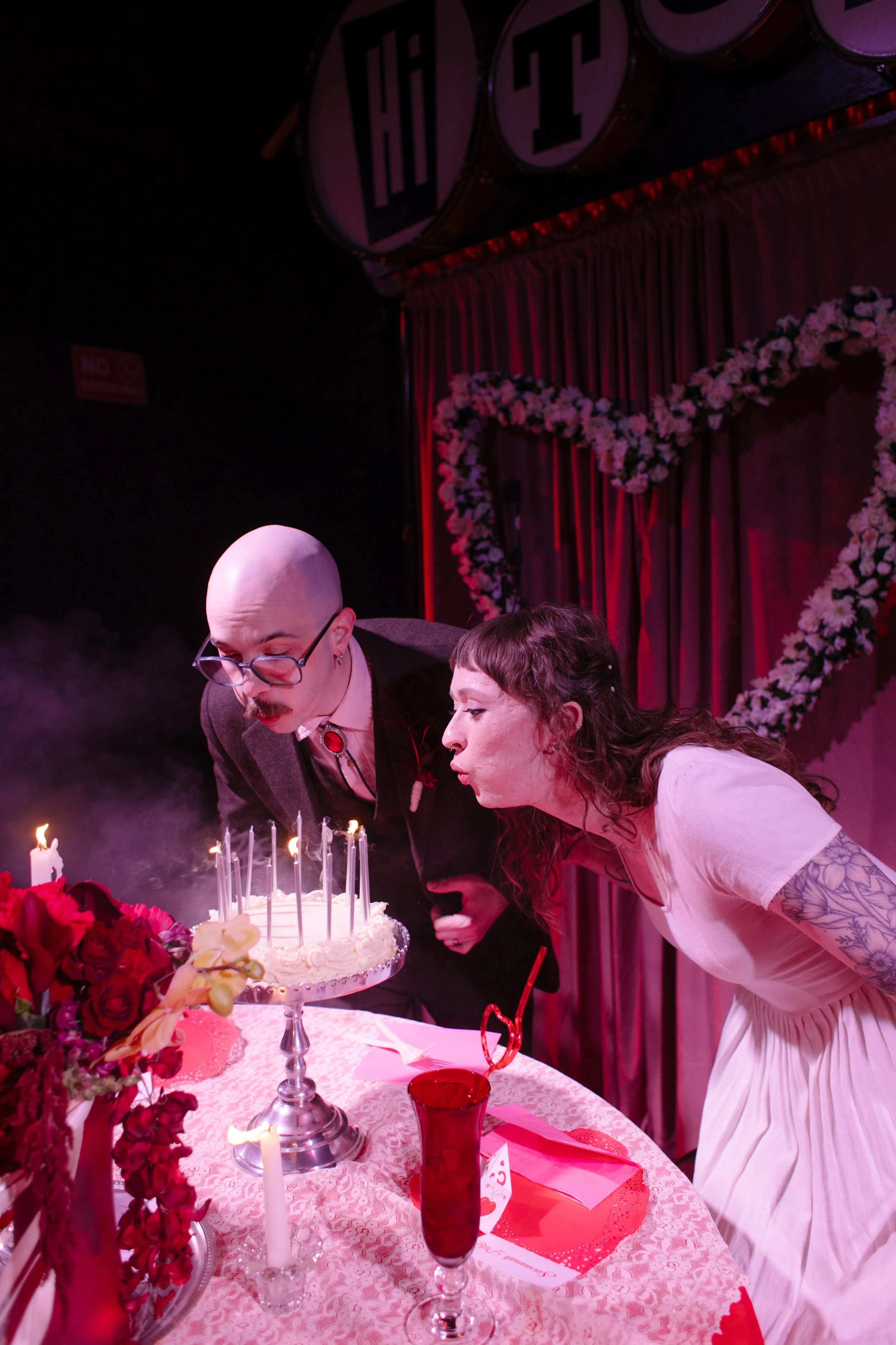Bride and groom blowing out candles on a small cake surrounded by red lighting and florals, capturing intimate and unconventional wedding details