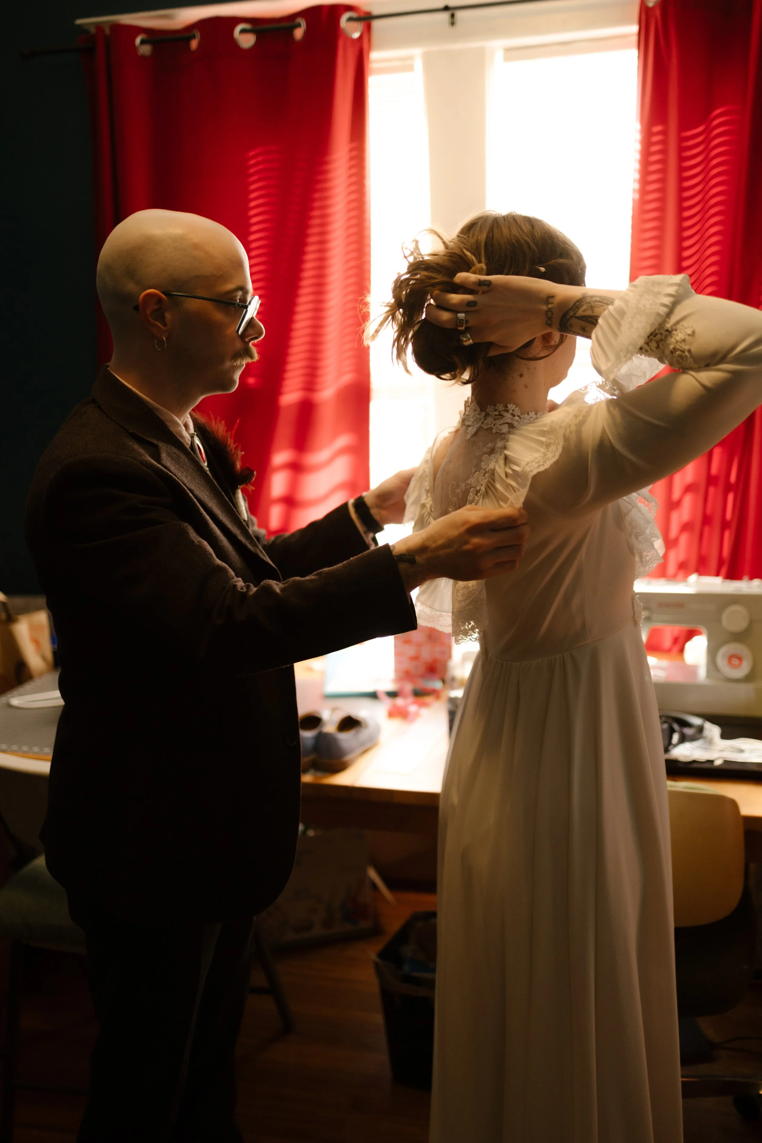 Groom helping button the bride’s dress in front of red curtains during wedding morning, showing quiet and meaningful moments