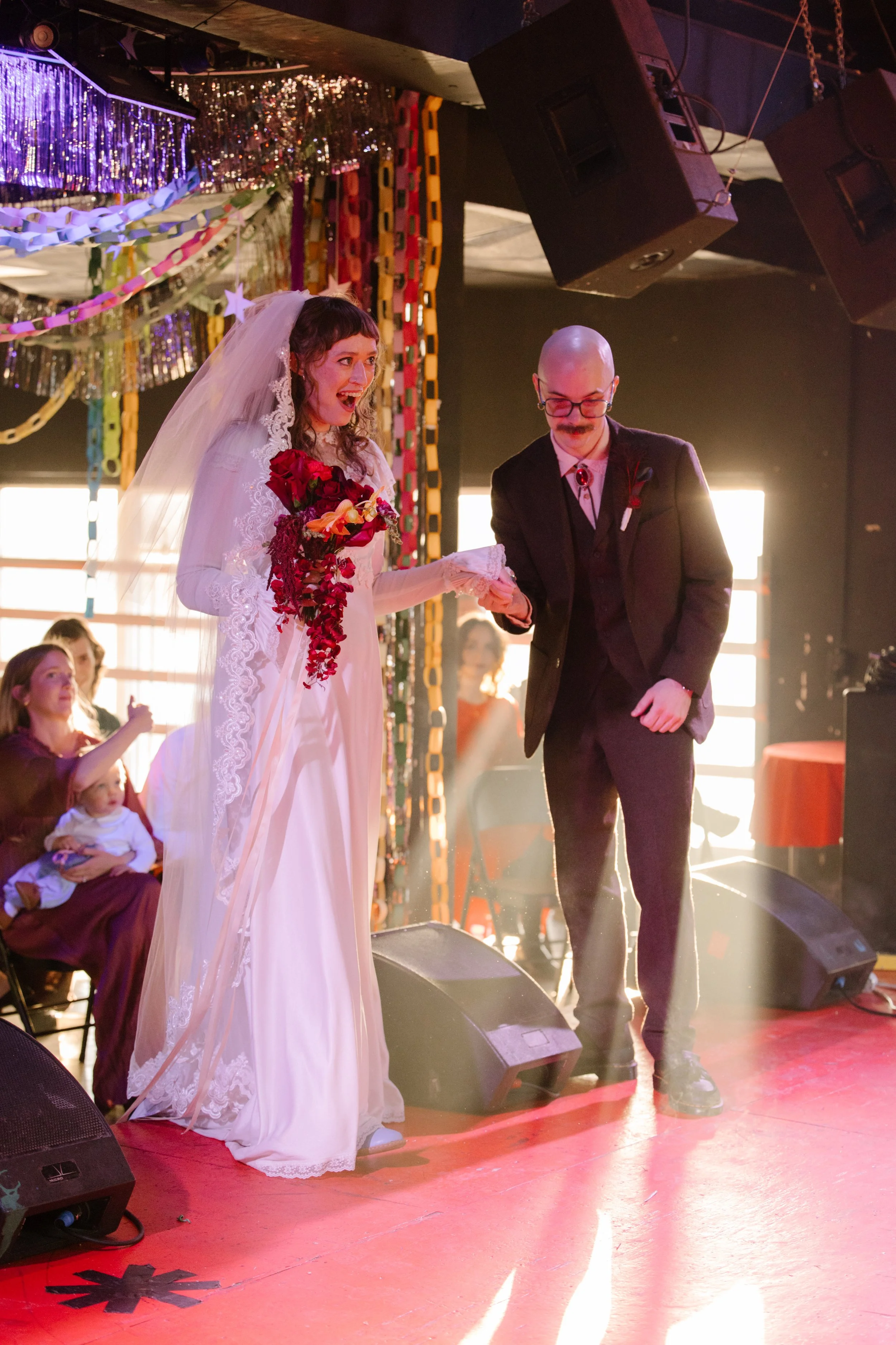 Bride stepping onto stage with groom during ceremony under colorful paper chains, capturing immersive and expressive unique wedding ideas