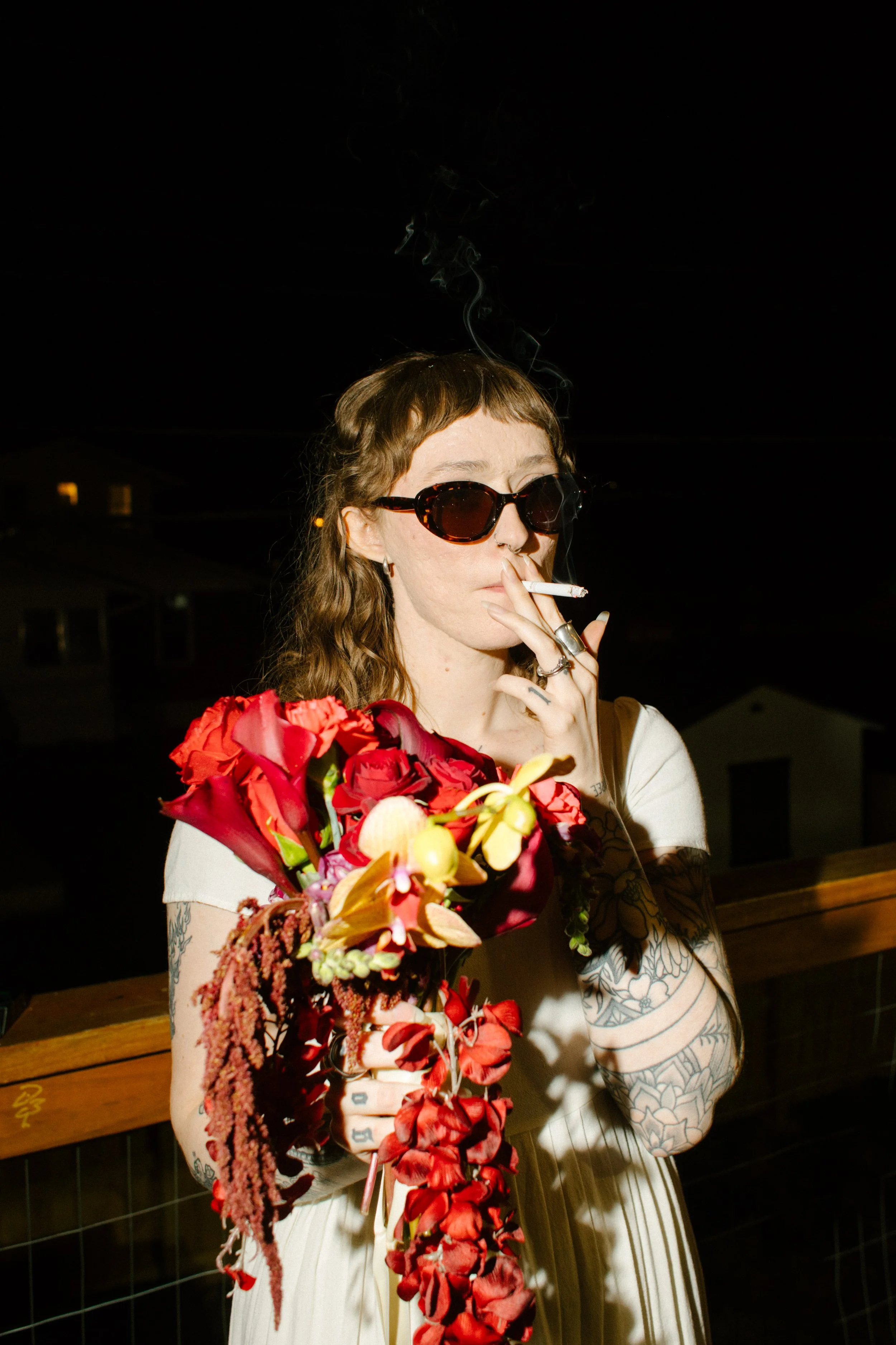 Bride wearing sunglasses and holding bold red bouquet while smoking, capturing edgy and unconventional unique wedding ideas