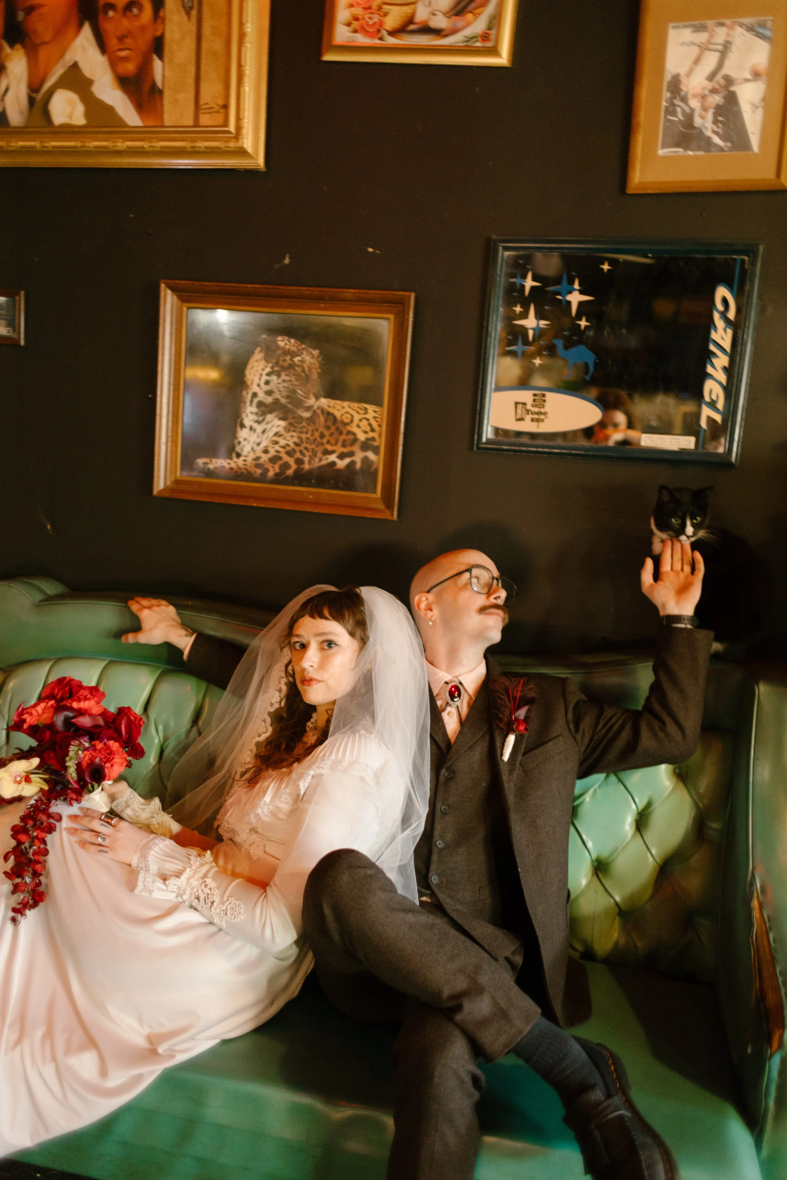 Bride and groom posing on green velvet couch with eclectic framed art and a cat, showcasing editorial and personality-driven unique wedding ideas