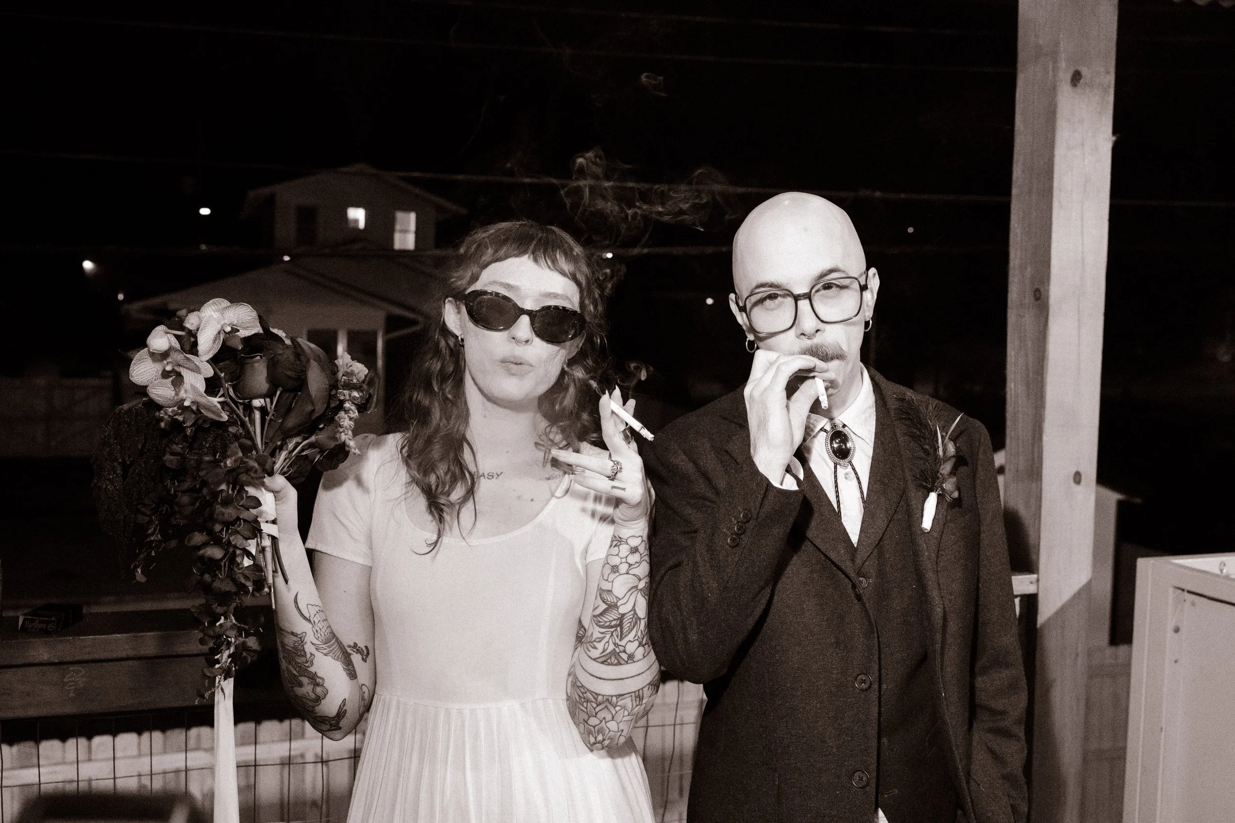 Black and white portrait of bride and groom smoking and holding bouquet at night, capturing edgy and documentary-style unique wedding ideas