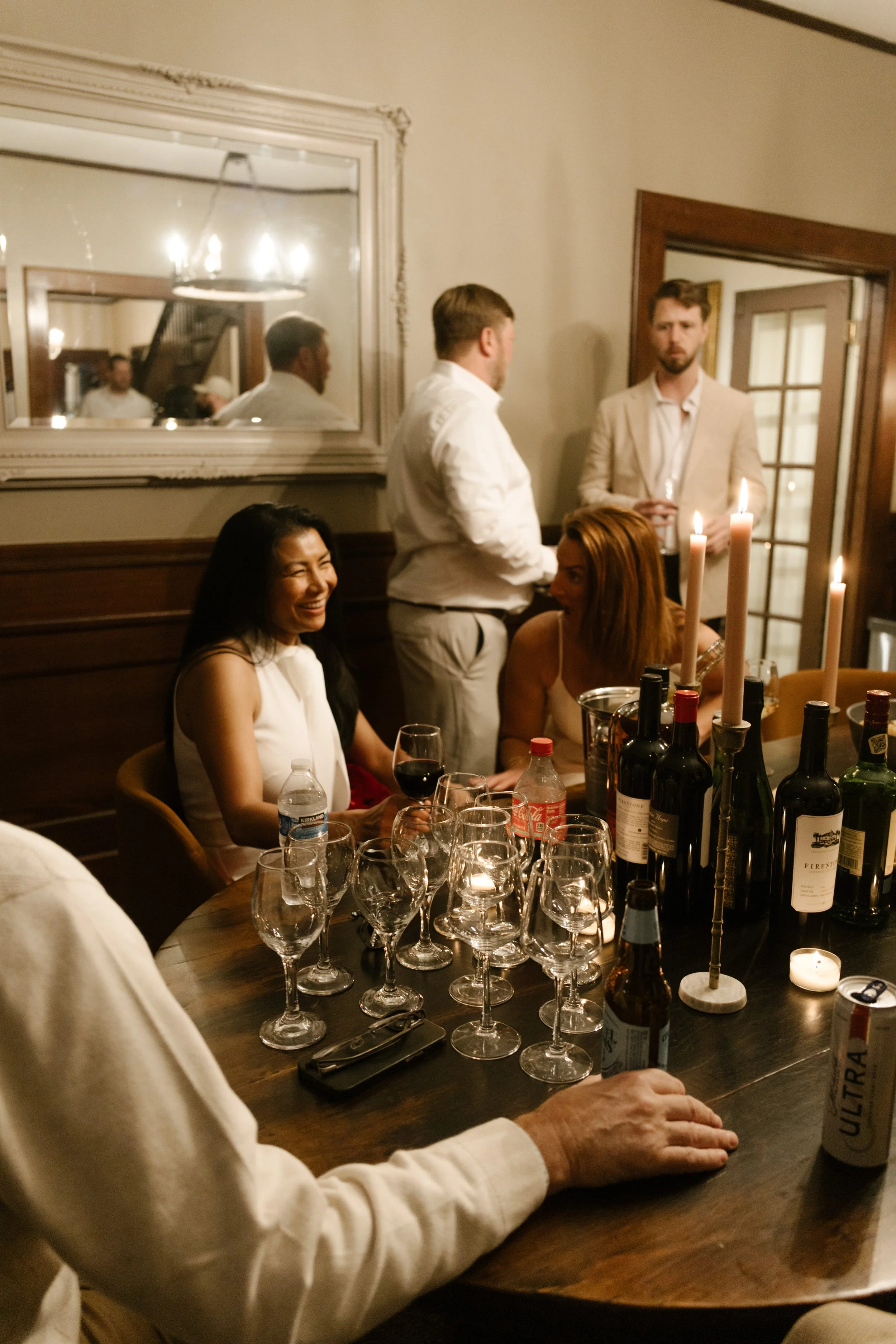 Intimate dinner scene with guests gathered around a candlelit table sharing drinks and conversation