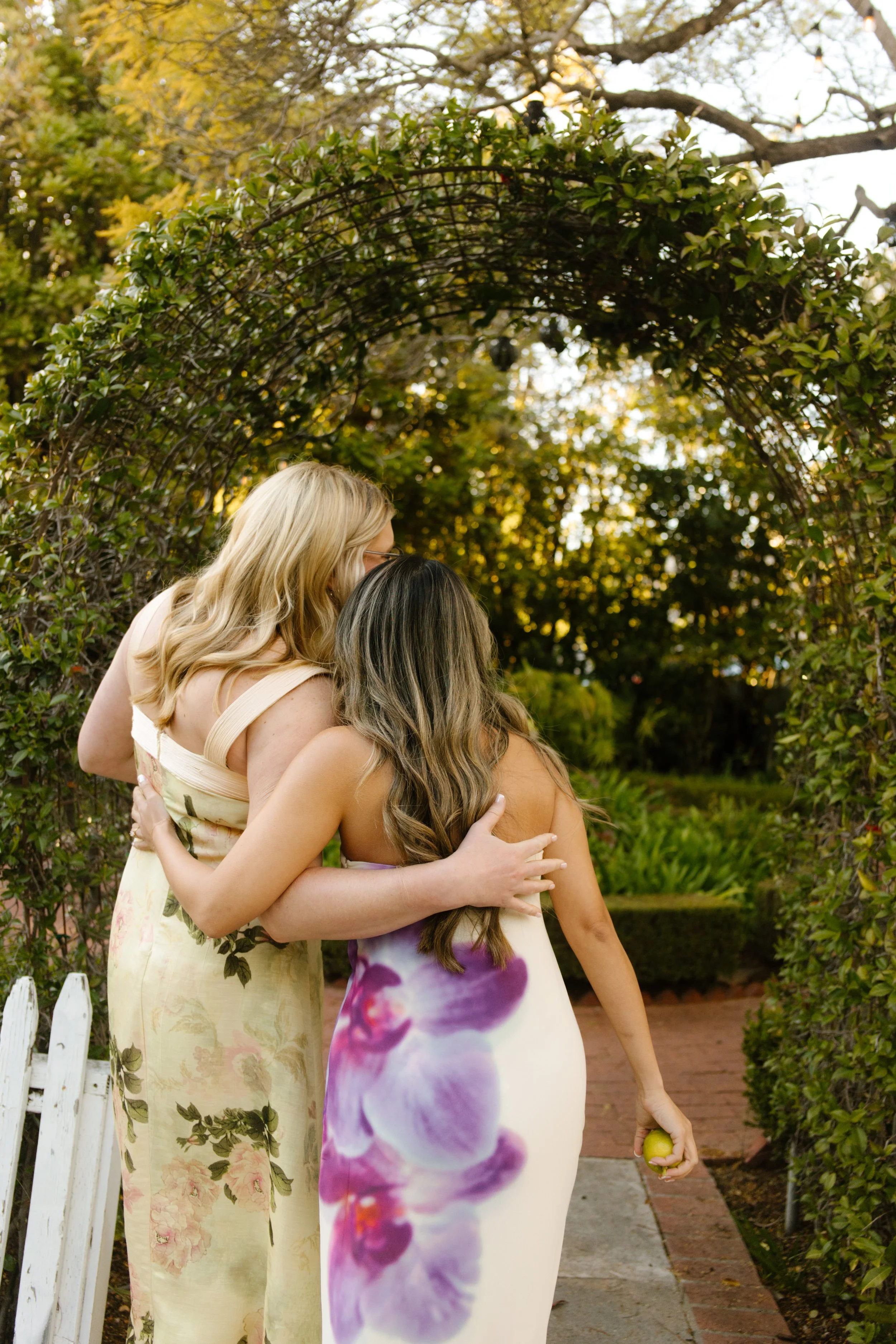 Two bridesmaids walking arm in arm under a garden archway holding a lemon during a wedding in Santa Barbara