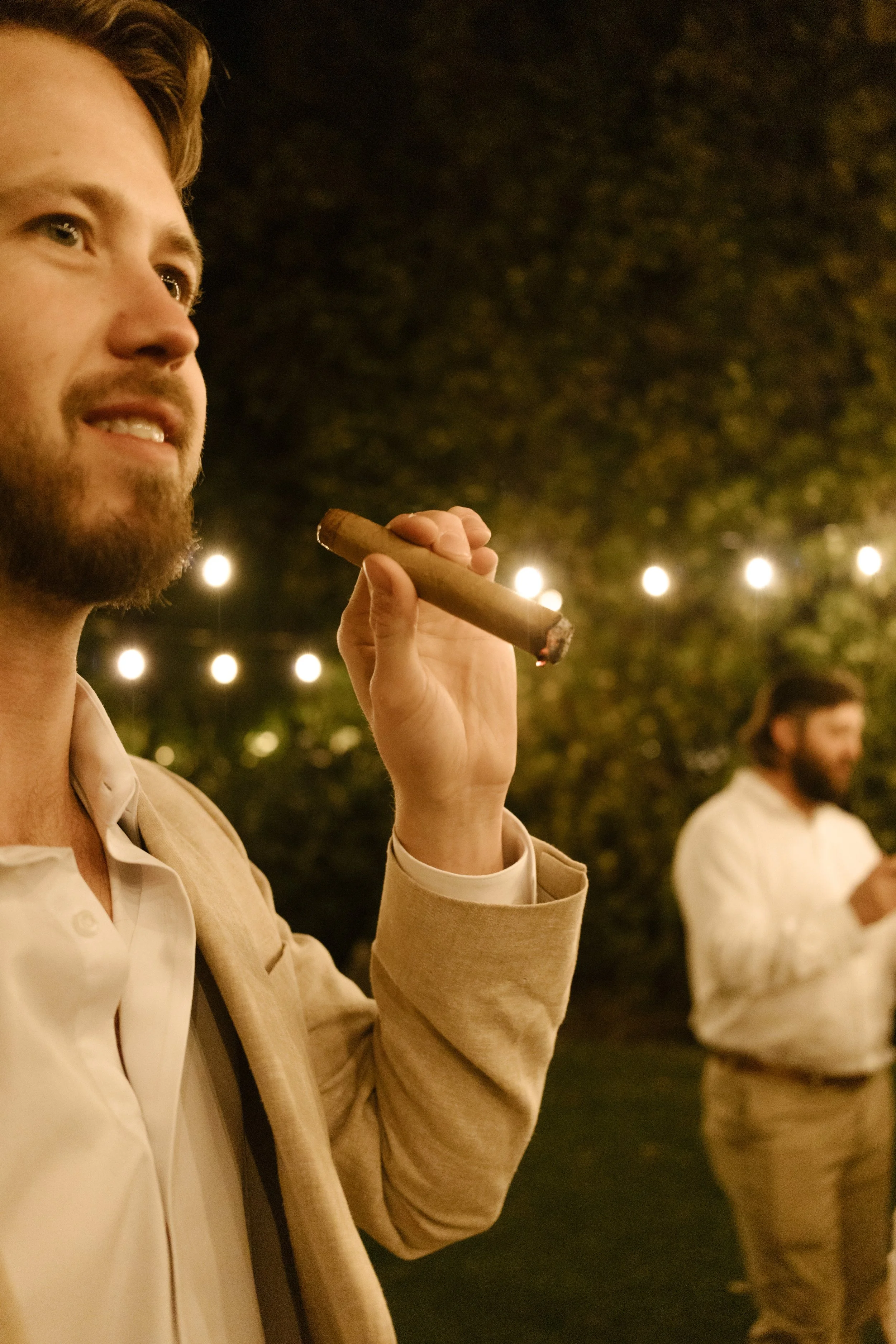 Groom smoking a cigar under string lights during a relaxed backyard moment at a wedding in Santa Barbara