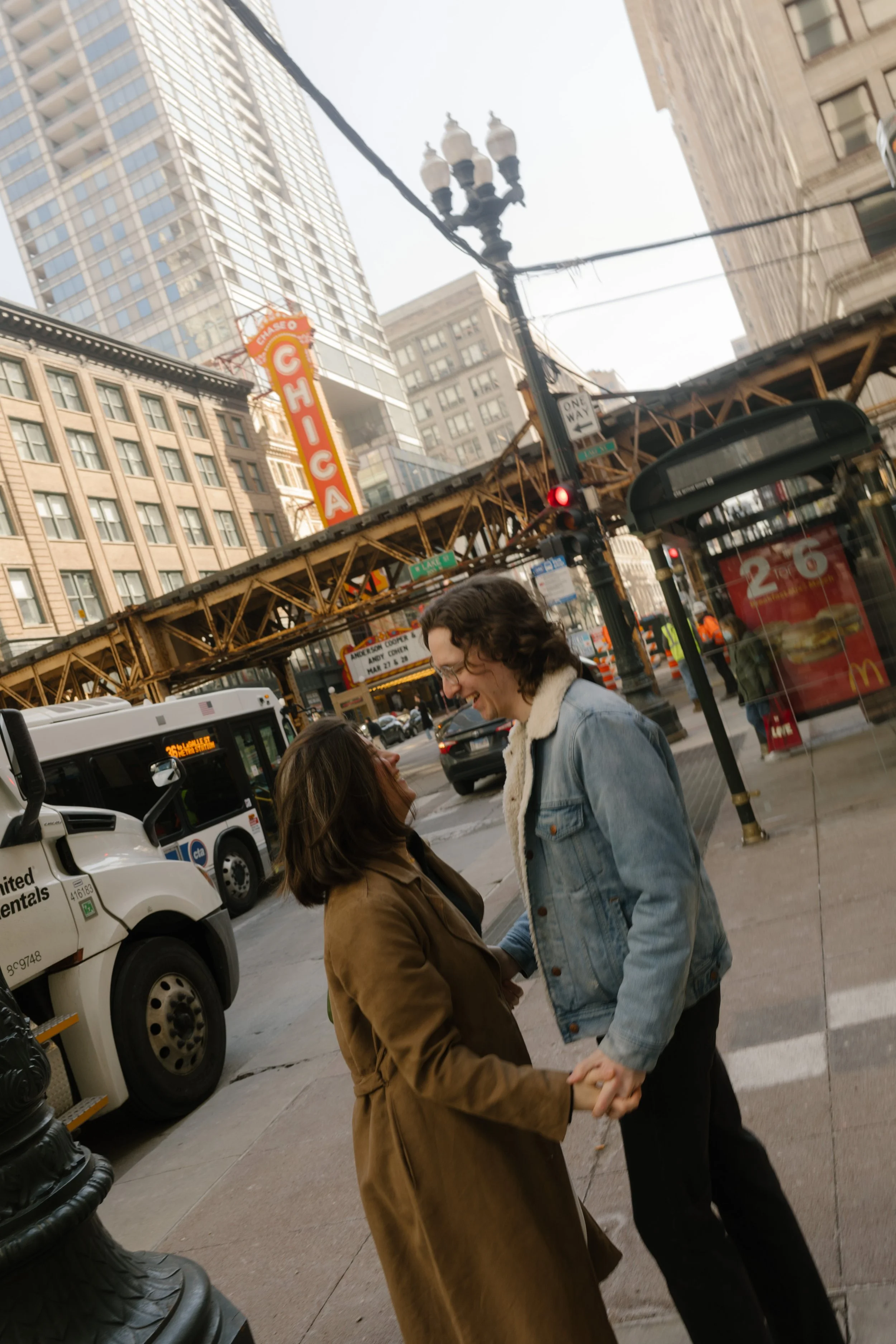 Couple laughing and holding hands on a busy downtown Chicago street beneath the elevated train tracks during engagement photos in Chicago