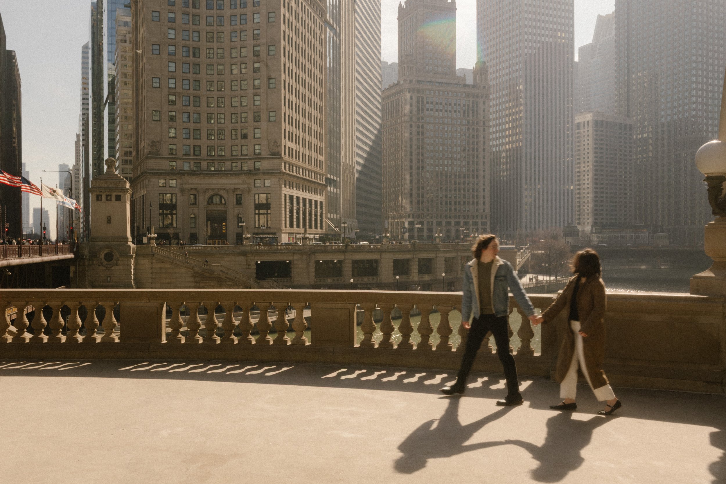Couple walking hand in hand across a bridge overlooking the Chicago River with skyline views during engagement photos in Chicago