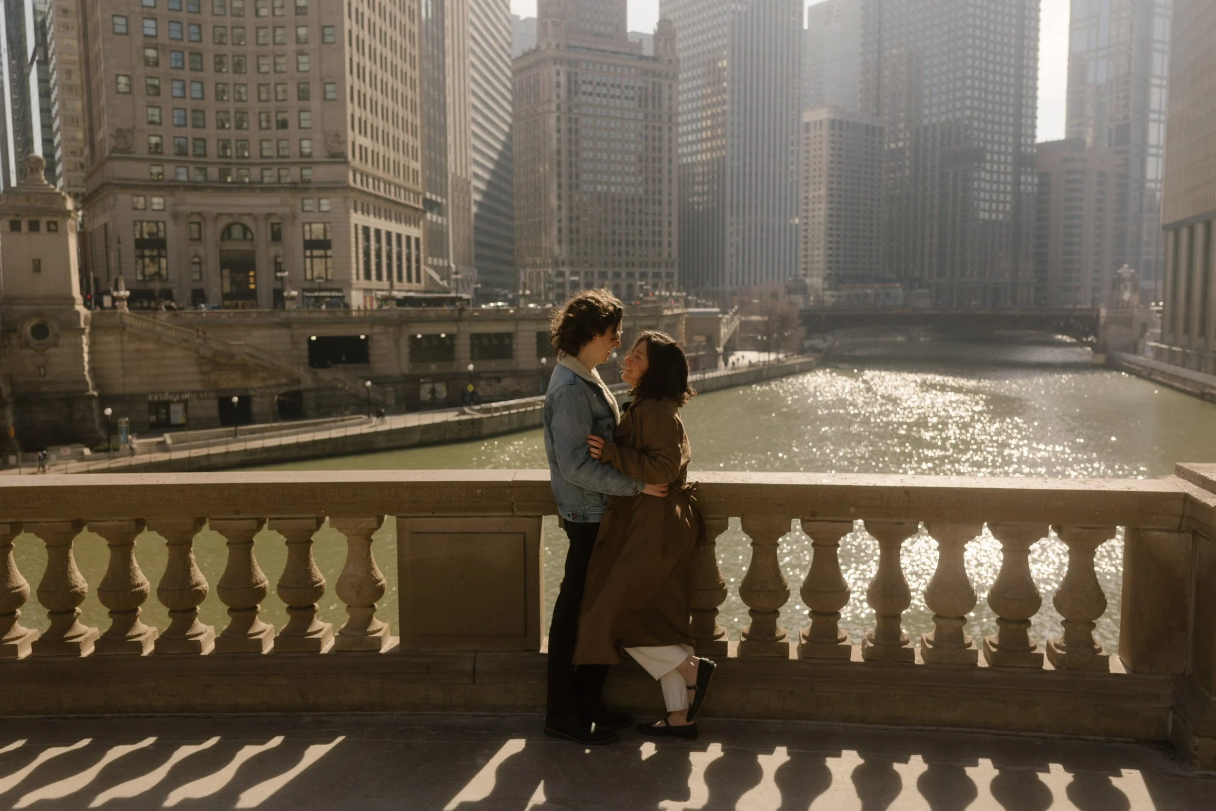 Couple standing close together on a stone bridge overlooking the Chicago River with sunlight reflecting on the water during engagement photos in Chicago