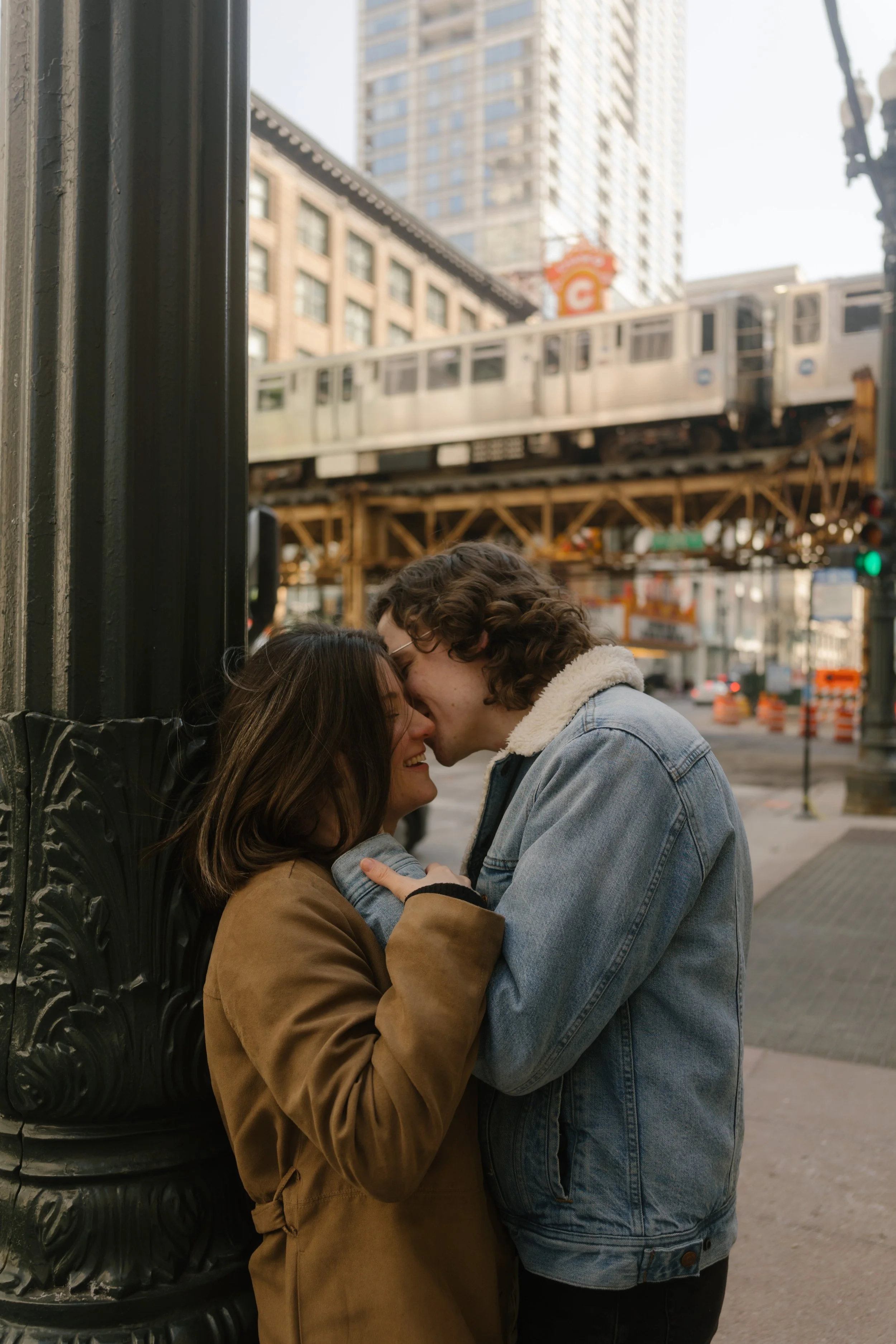 Intimate moment of couple leaning into each other near a city streetlight with a train passing overhead in downtown Chicago during engagement photos in Chicago