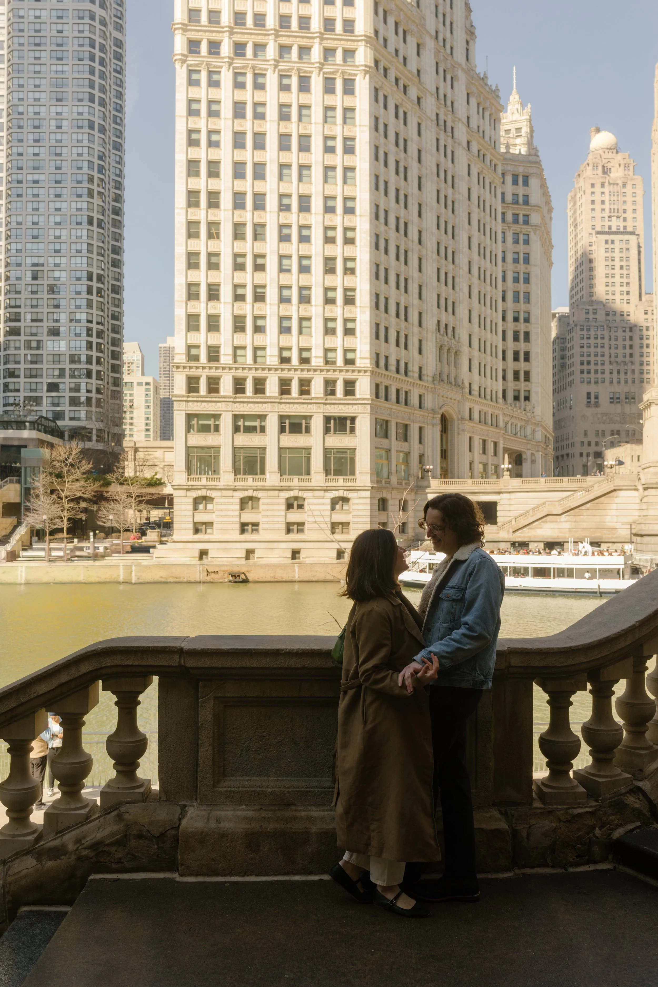 Couple holding hands on stone steps along the Chicago River with historic buildings rising behind them
